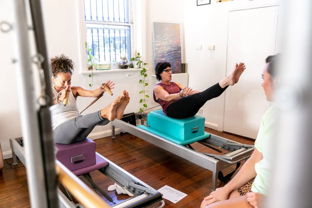 Two women exercising on Pilates reformers with a trainer observing in a bright studio.