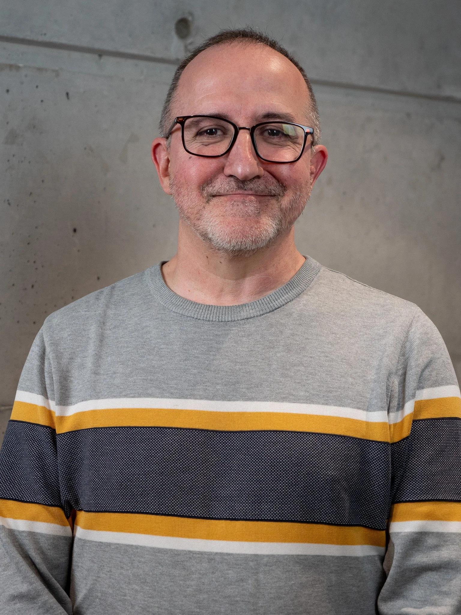 Un homme souriant portant des lunettes, habillé d'un pull gris à rayures jaunes, blanches et noires, se tenant devant un mur en béton.