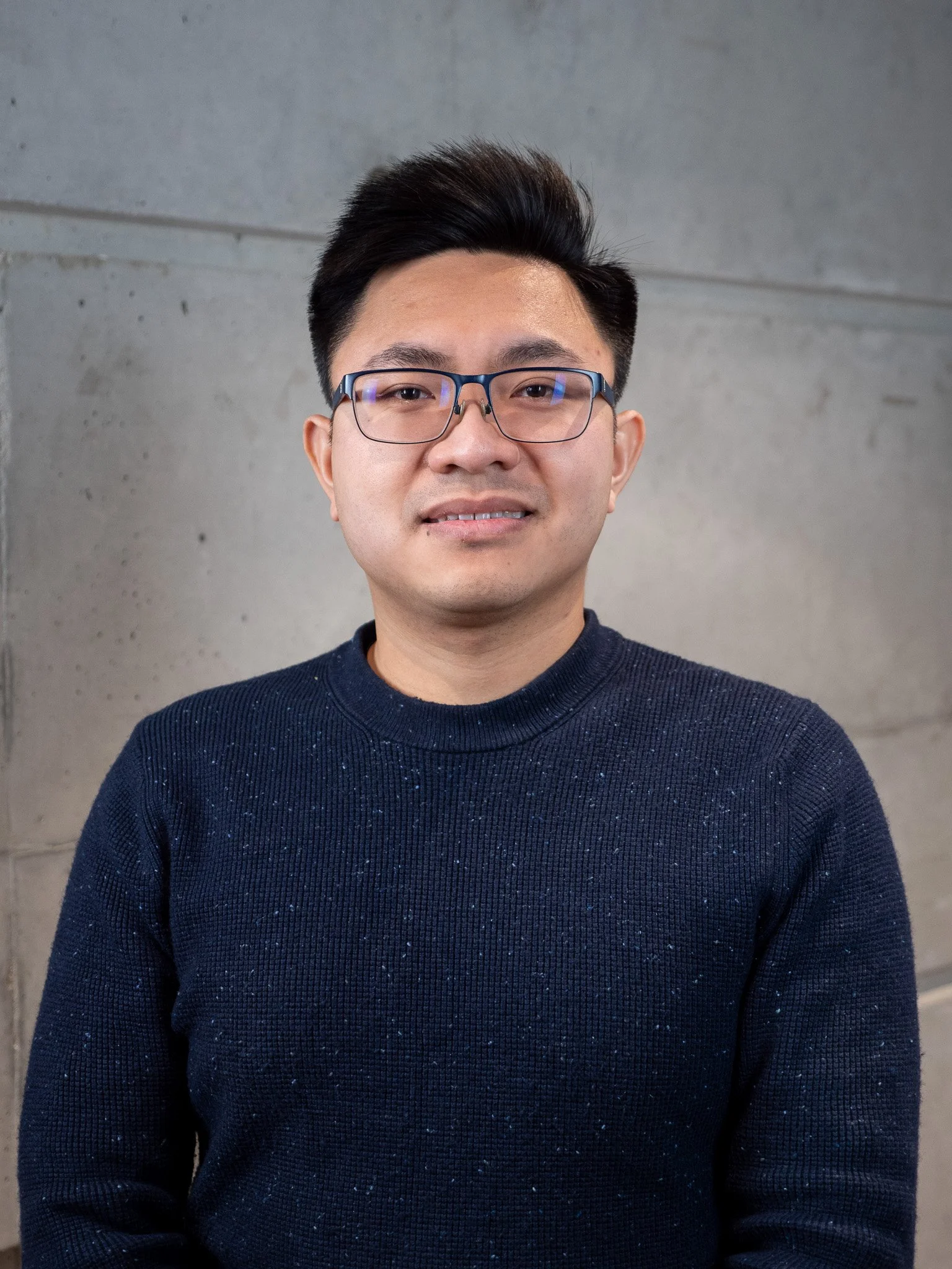 Jeune homme asiatique avec des lunettes et un pull bleu foncé, souriant, en face d'un mur en béton.