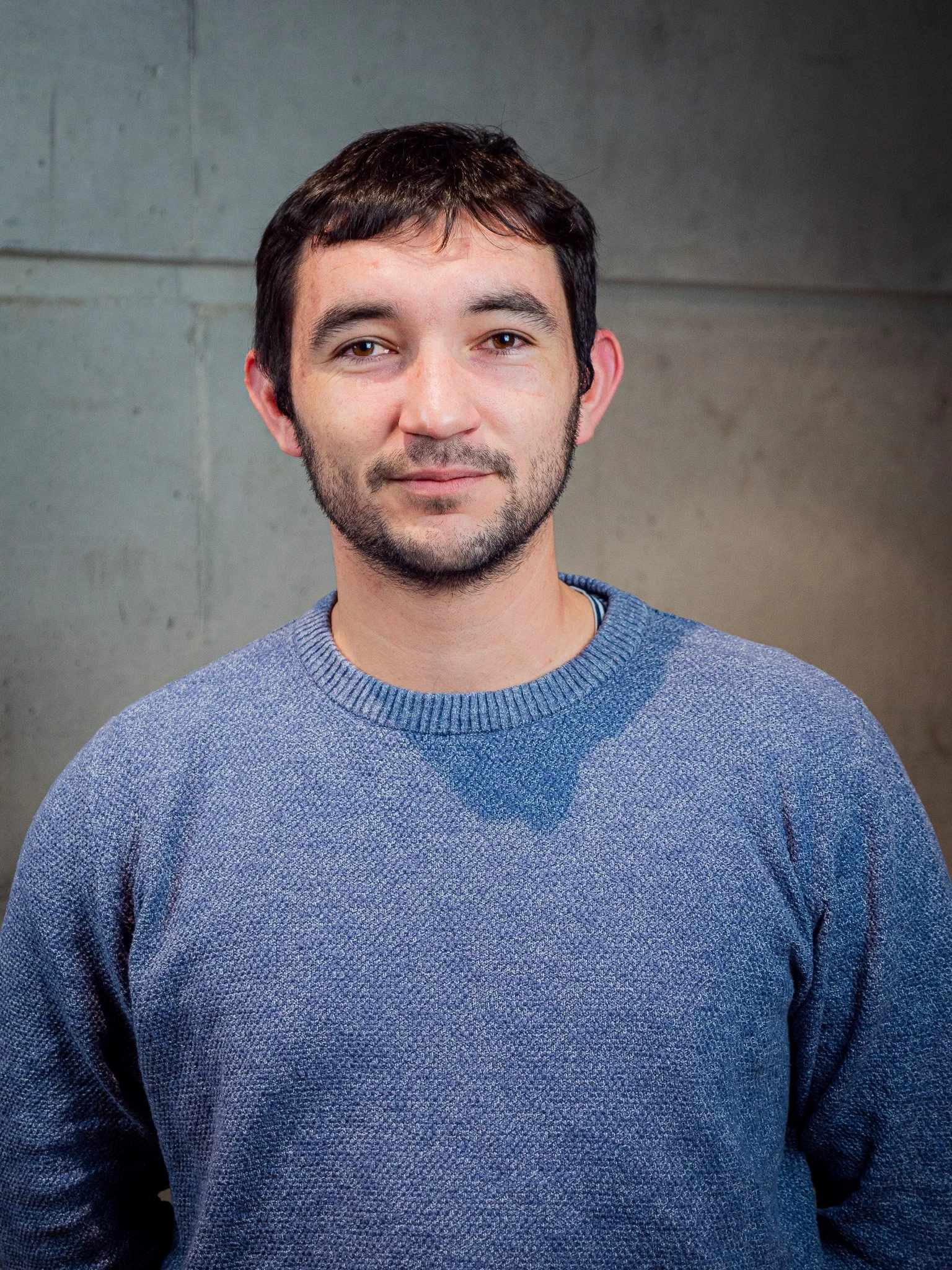 Homme portant un pull bleu clair, souriant légèrement, avec un mur en béton en arrière-plan.