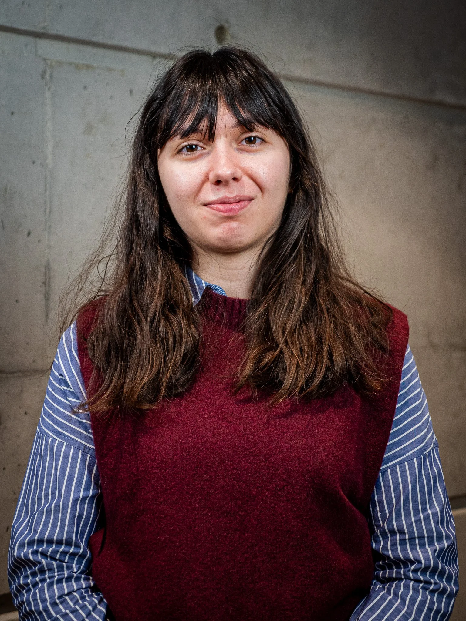 Une jeune femme à cheveux bruns, portant une chemise à rayures bleues sous un pull marron, se tient devant un mur en béton.