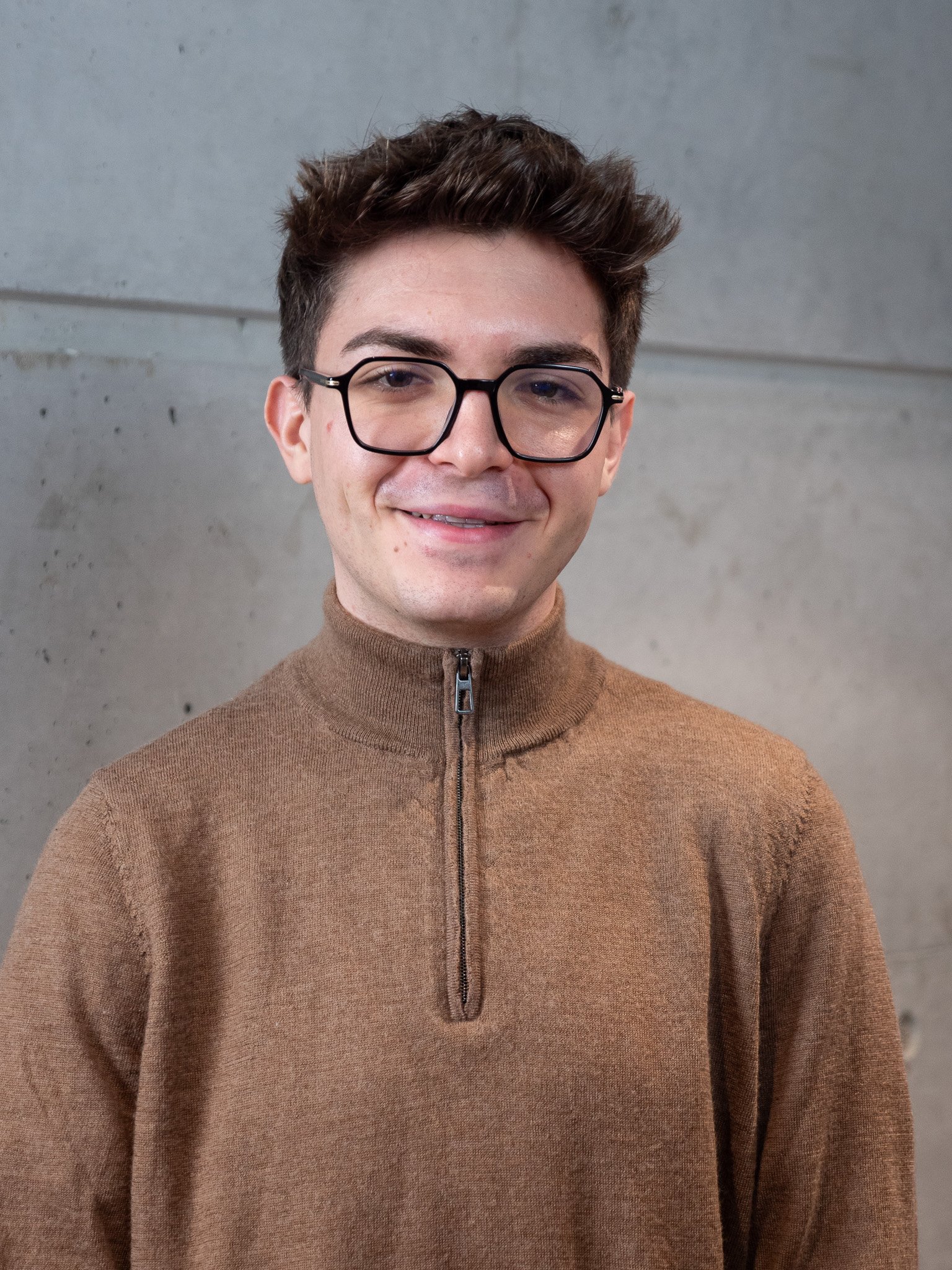 Jeune homme souriant portant un col roulé blanc devant un mur en béton.