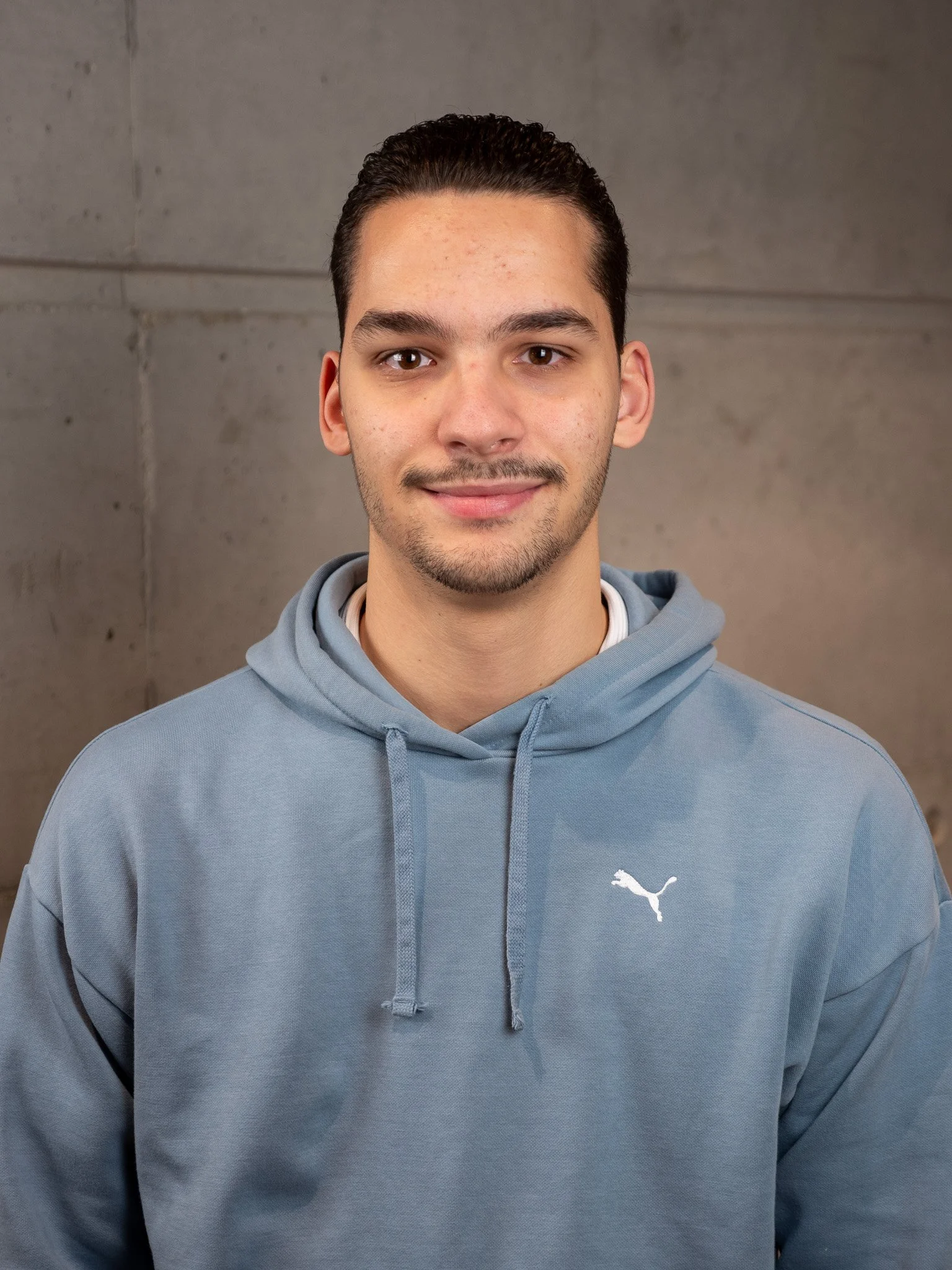 Jeune homme souriant portant un t-shirt beige avec le logo 'Jack & Jones' en blanc et bleu, debout contre un mur en béton gris.