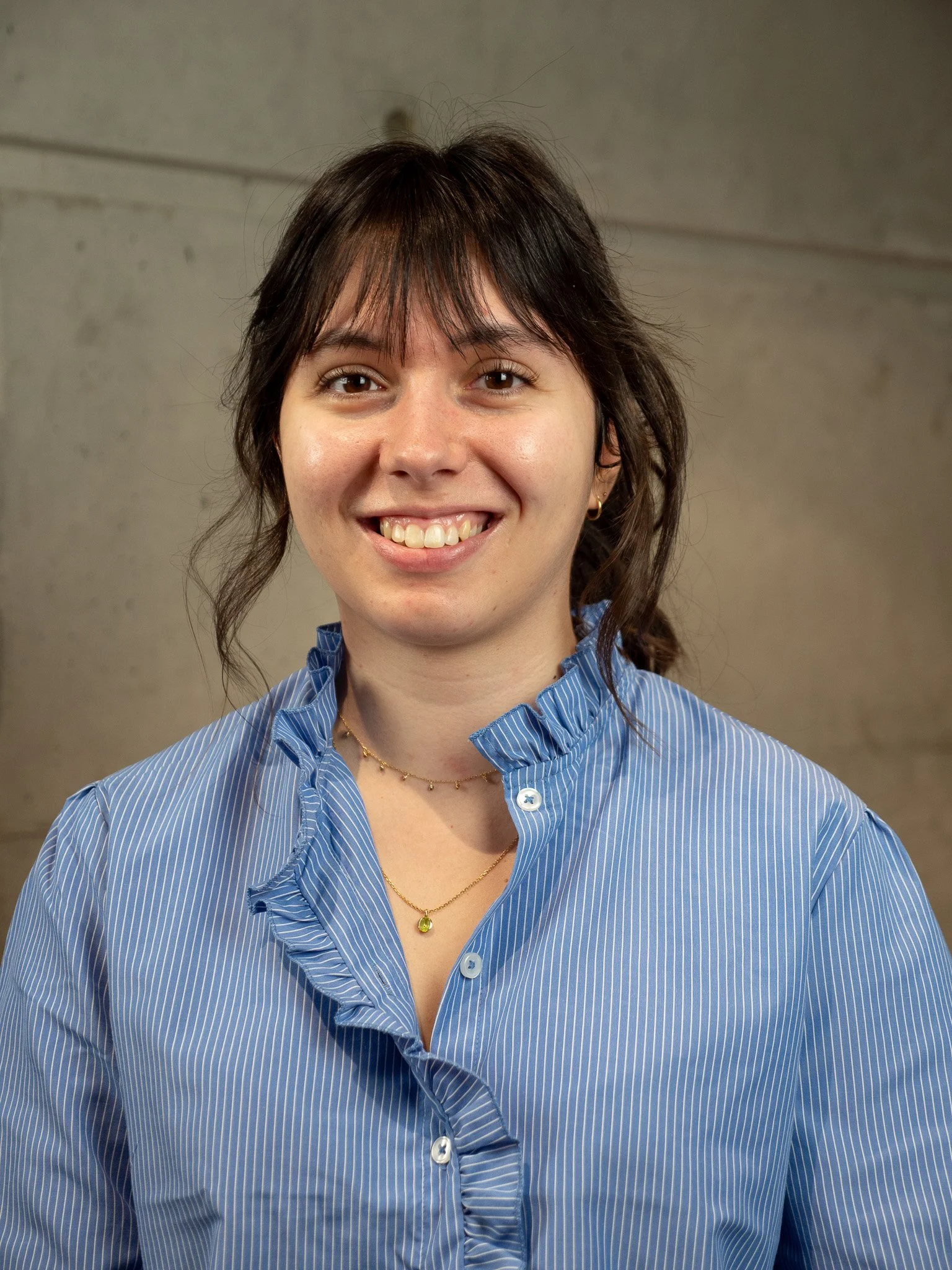 Une jeune femme à cheveux bruns, portant une chemise à rayures bleues sous un pull marron, se tient devant un mur en béton.