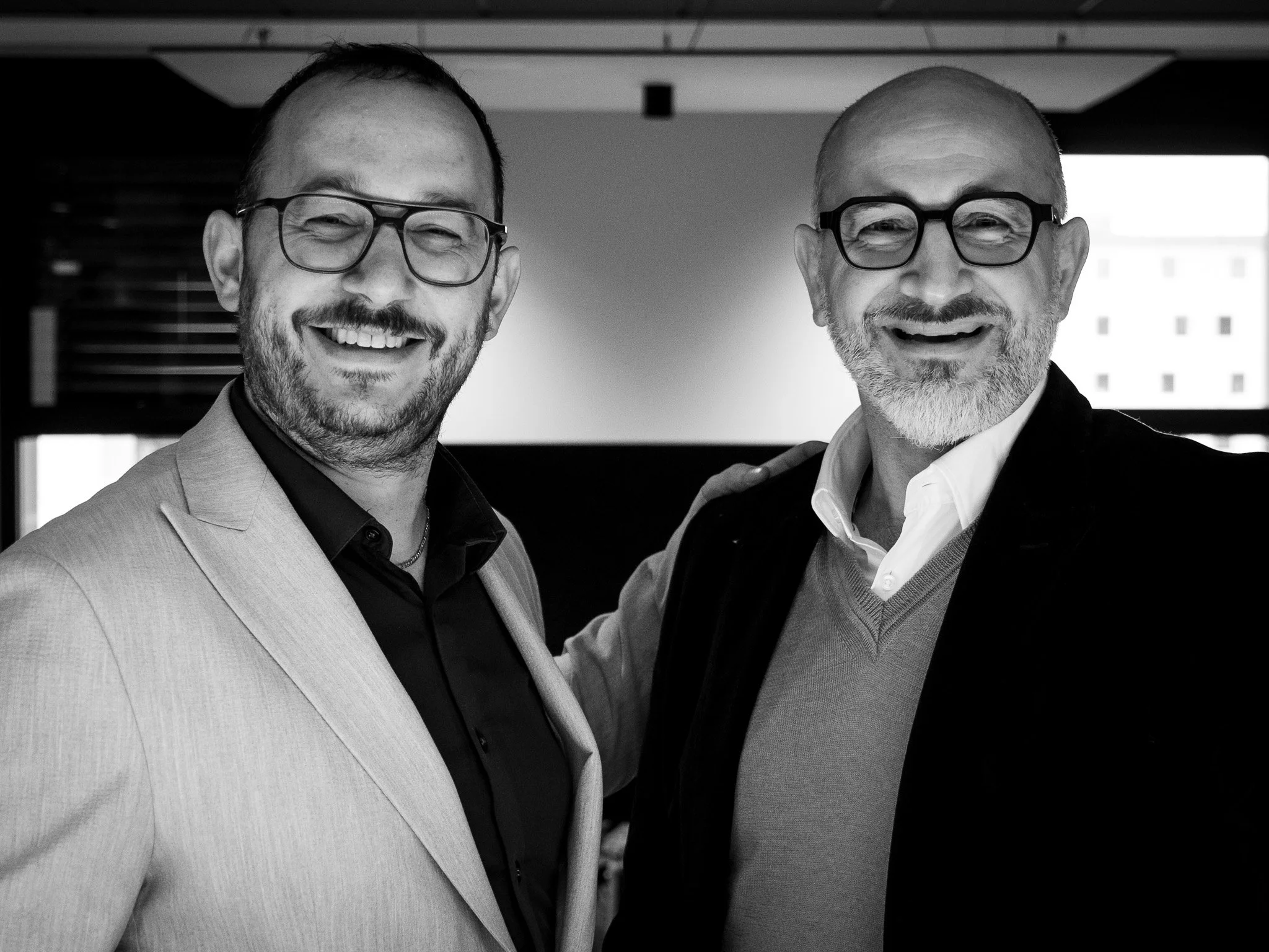 Deux hommes souriants portant des lunettes, l'un en costume clair et l'autre en veste sombre, posant ensemble à l'intérieur.