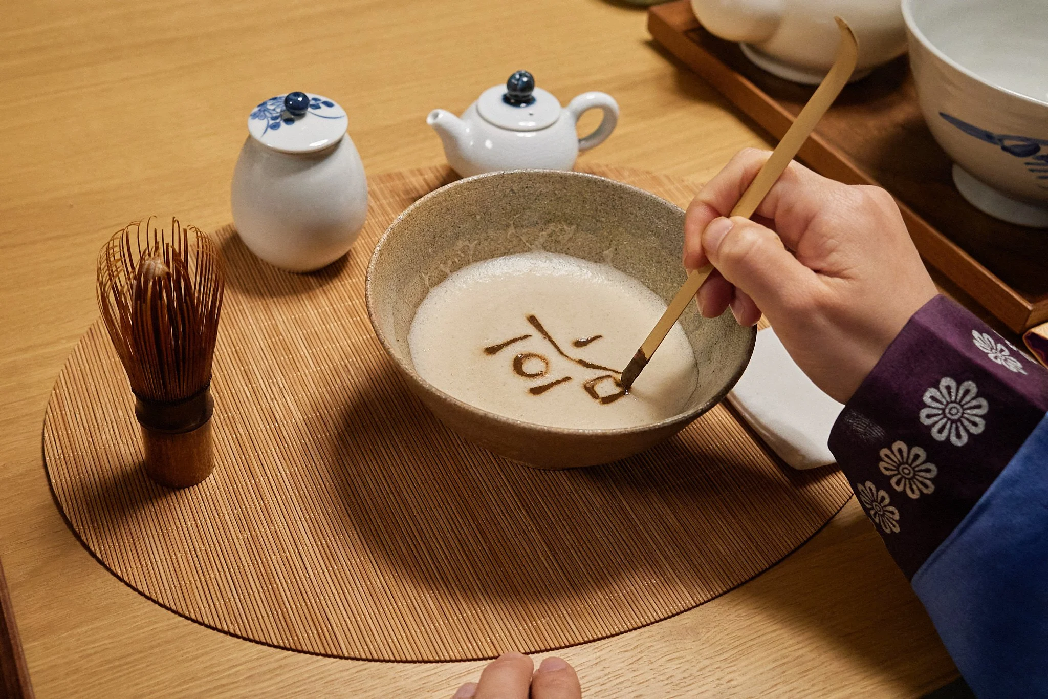 Person decorating a bowl of rice porridge with a dark sauce using a chopstick, surrounded by teapot and containers on a wooden table.