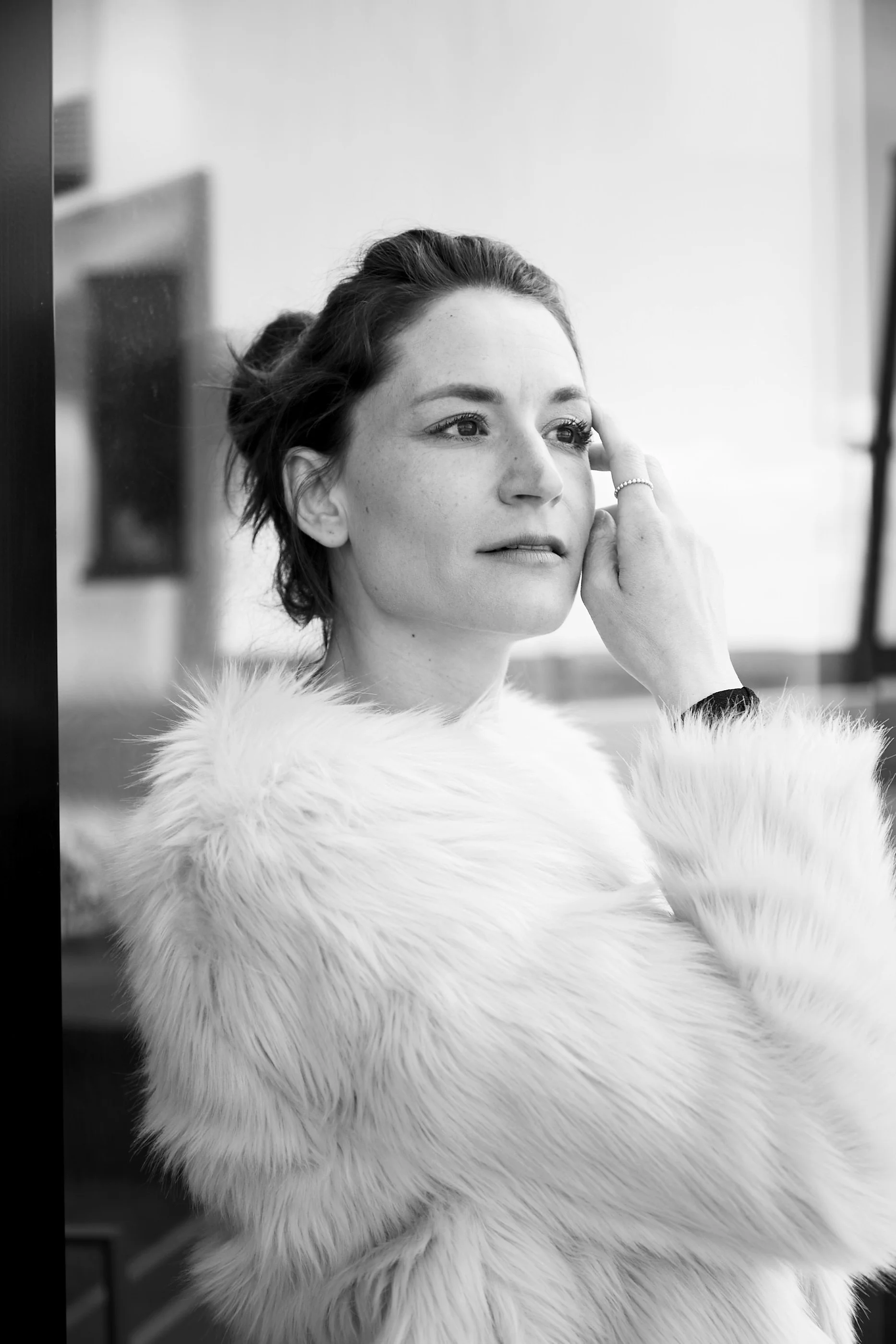 A woman with short dark hair and light skin, wearing a fluffy fur coat, looks thoughtfully into a mirror with her hand touching her cheek.