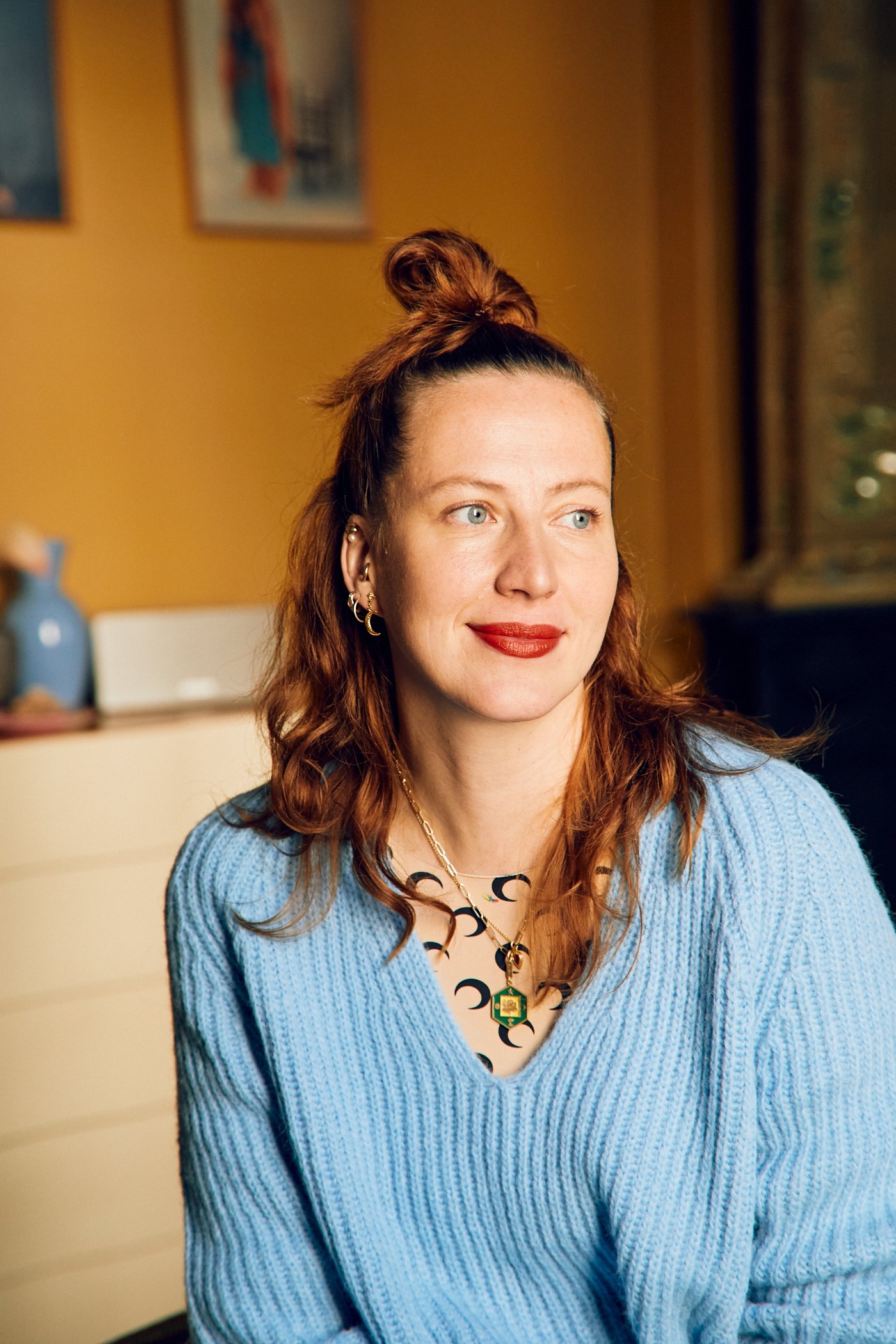 A woman with red hair styled in a messy bun, wearing a blue sweater, gold jewelry, and red lipstick, sitting in a room with warm-colored walls and framed artwork in the background.