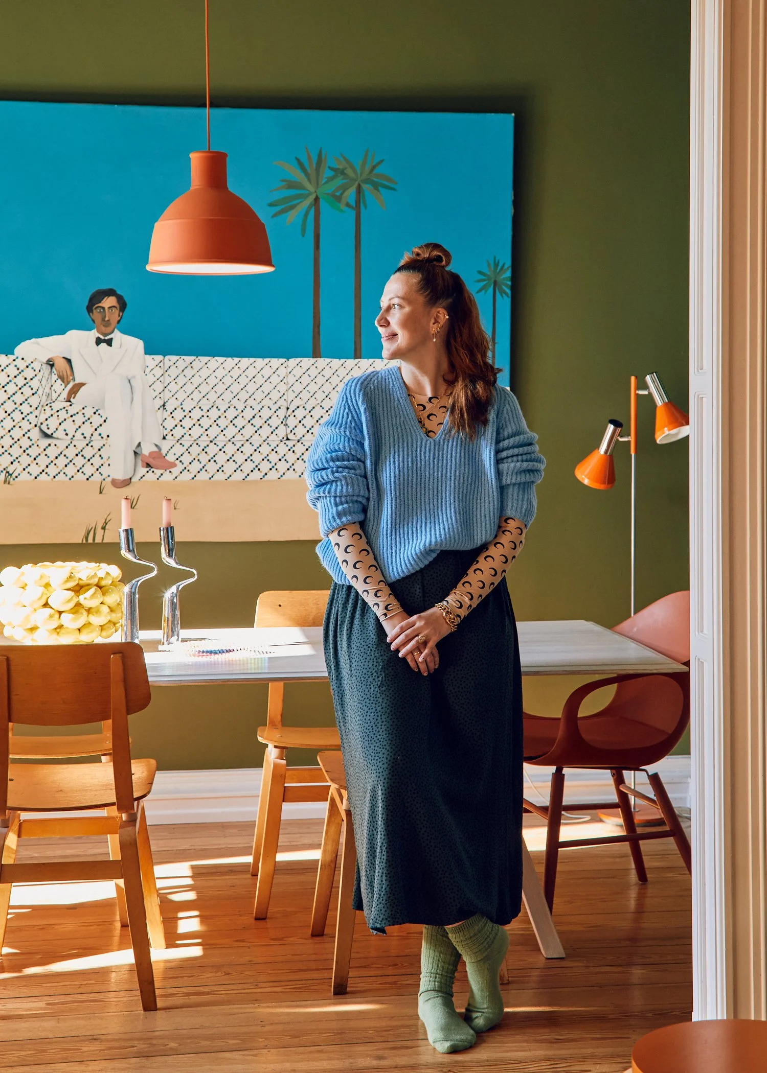 A woman in a blue sweater and patterned long-sleeve shirt standing in a decorated dining room, with a painting of a man in a white suit and palm trees on the wall behind her.