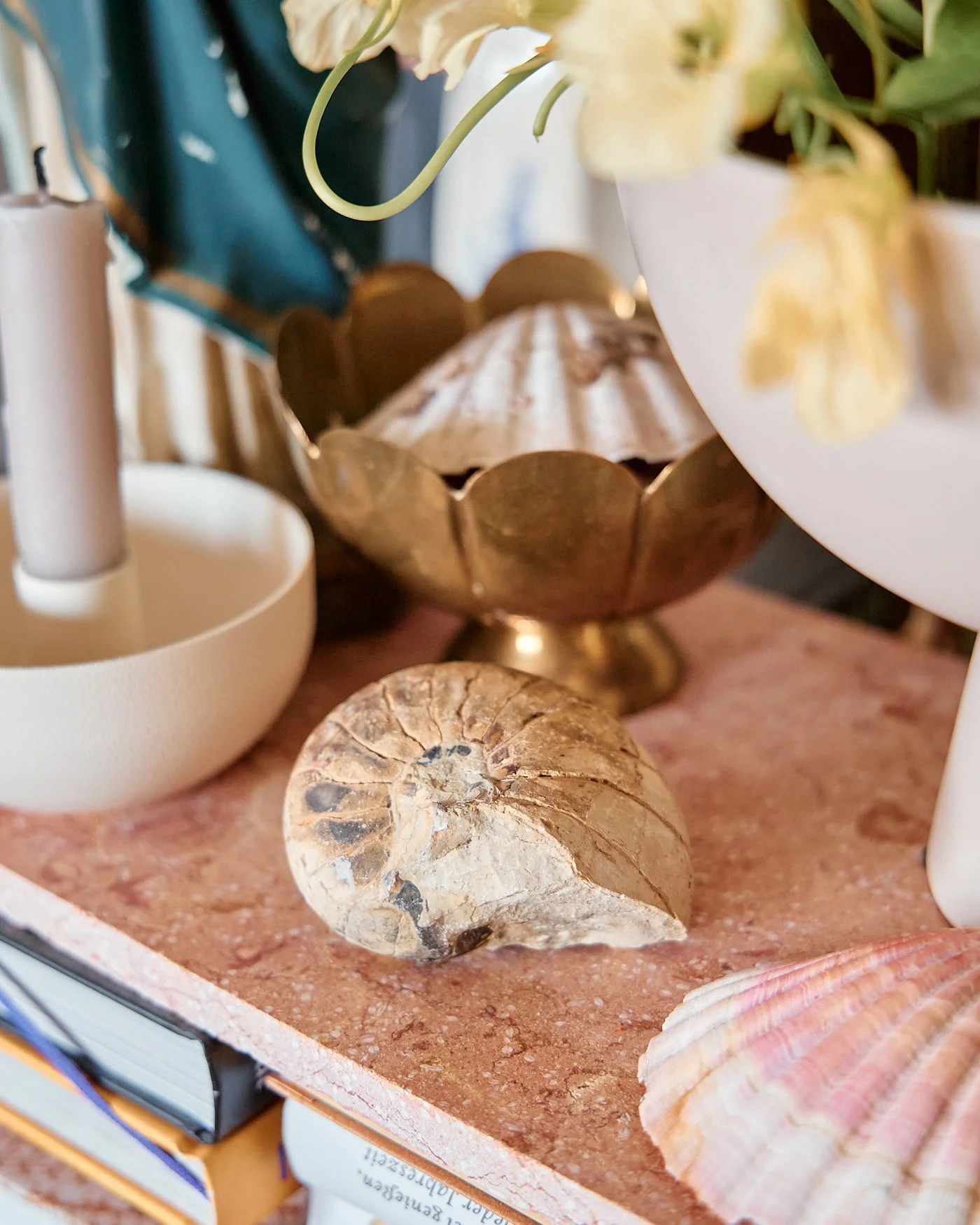 Close-up of a decorative arrangement on a pink stone surface, including a fossilized shell, a brass bowl, a white candlestick with a candle, and vases with flowers.