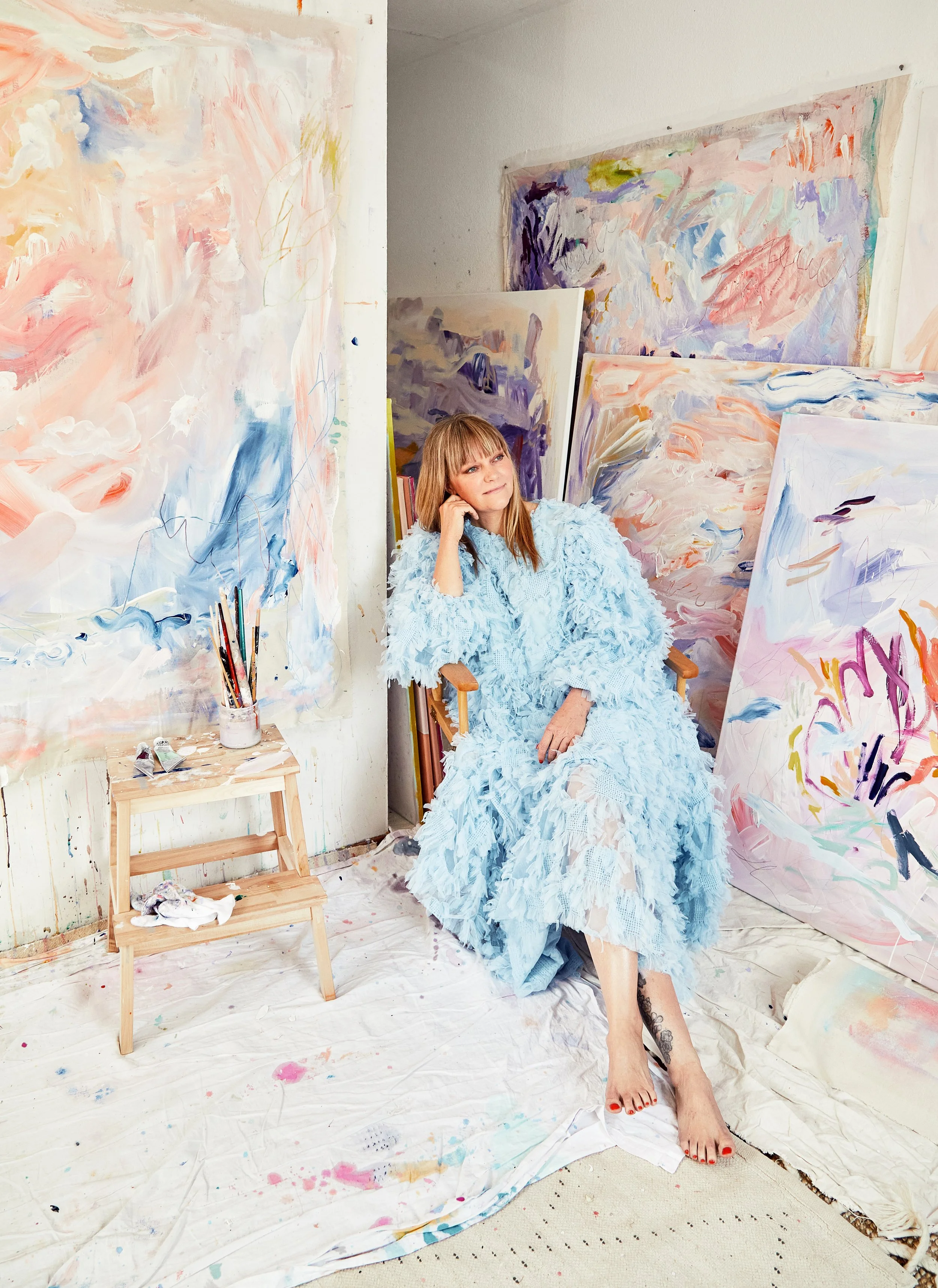 Woman sitting in a colorful abstract art studio surrounded by large paintings and art supplies.