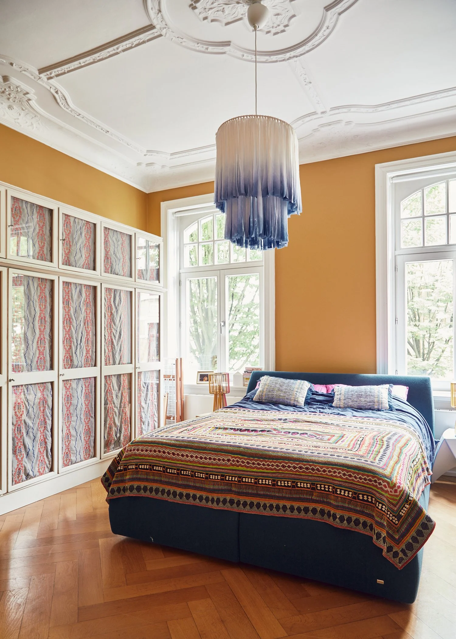 Bright bedroom with large windows, mustard-colored wall, and ornate white ceiling. A modern blue bed with a patterned quilt and multiple pillows. Decorative hanging light fixture with blue ombre fringe. White cabinet with patterned fabric behind glas