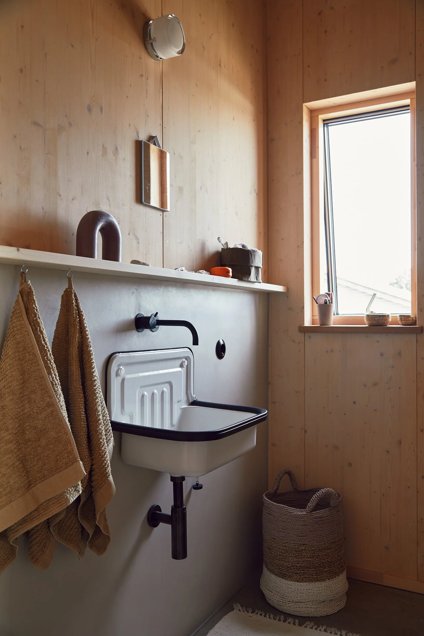 A minimalist laundry room with a small utility sink, beige towels hanging on the left, a wooden wall, a window letting in natural light, and a woven basket on the floor.