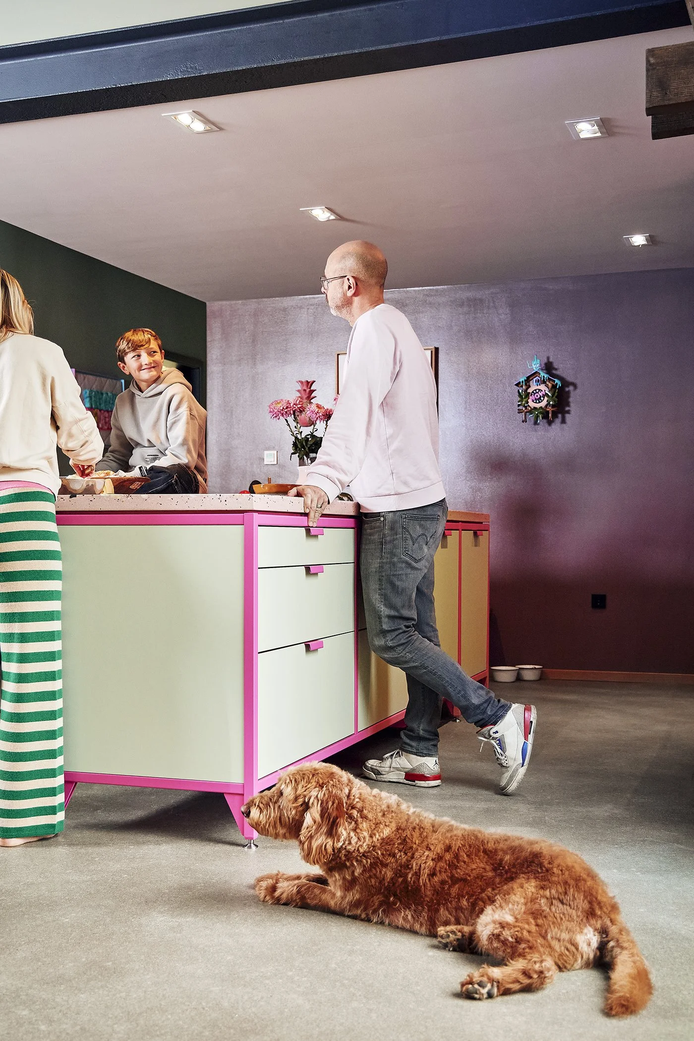 A scene in a kitchen with three people and a dog. A woman with striped pants is talking to a boy sitting at the counter. A man is standing and leaning on the kitchen counter, with a brown, curly-haired dog lying on the floor nearby.