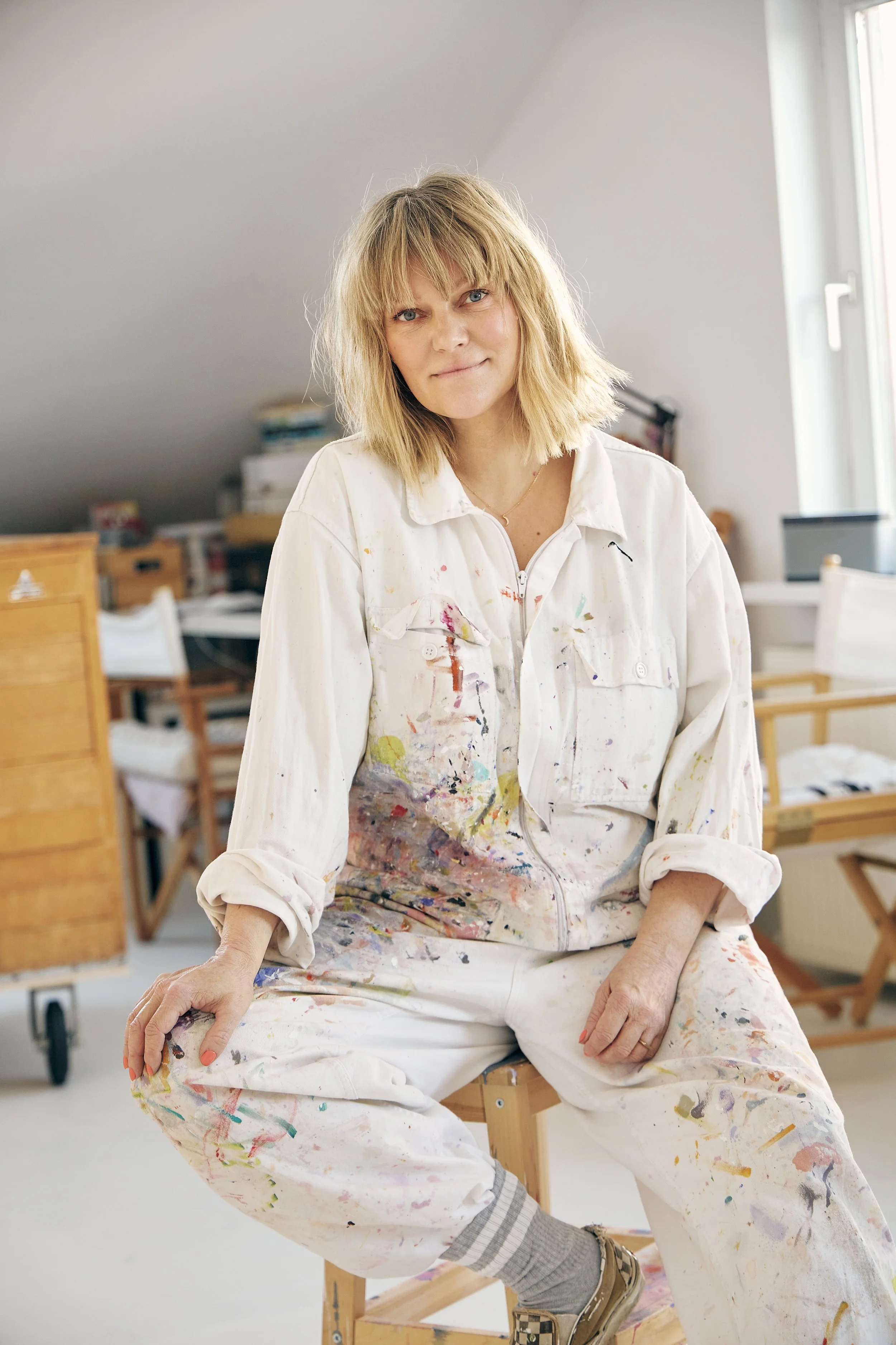 A woman with blonde hair and blue eyes, wearing a paint-splattered white jumpsuit, sitting on a wooden stool in an art studio with a neutral expression.
