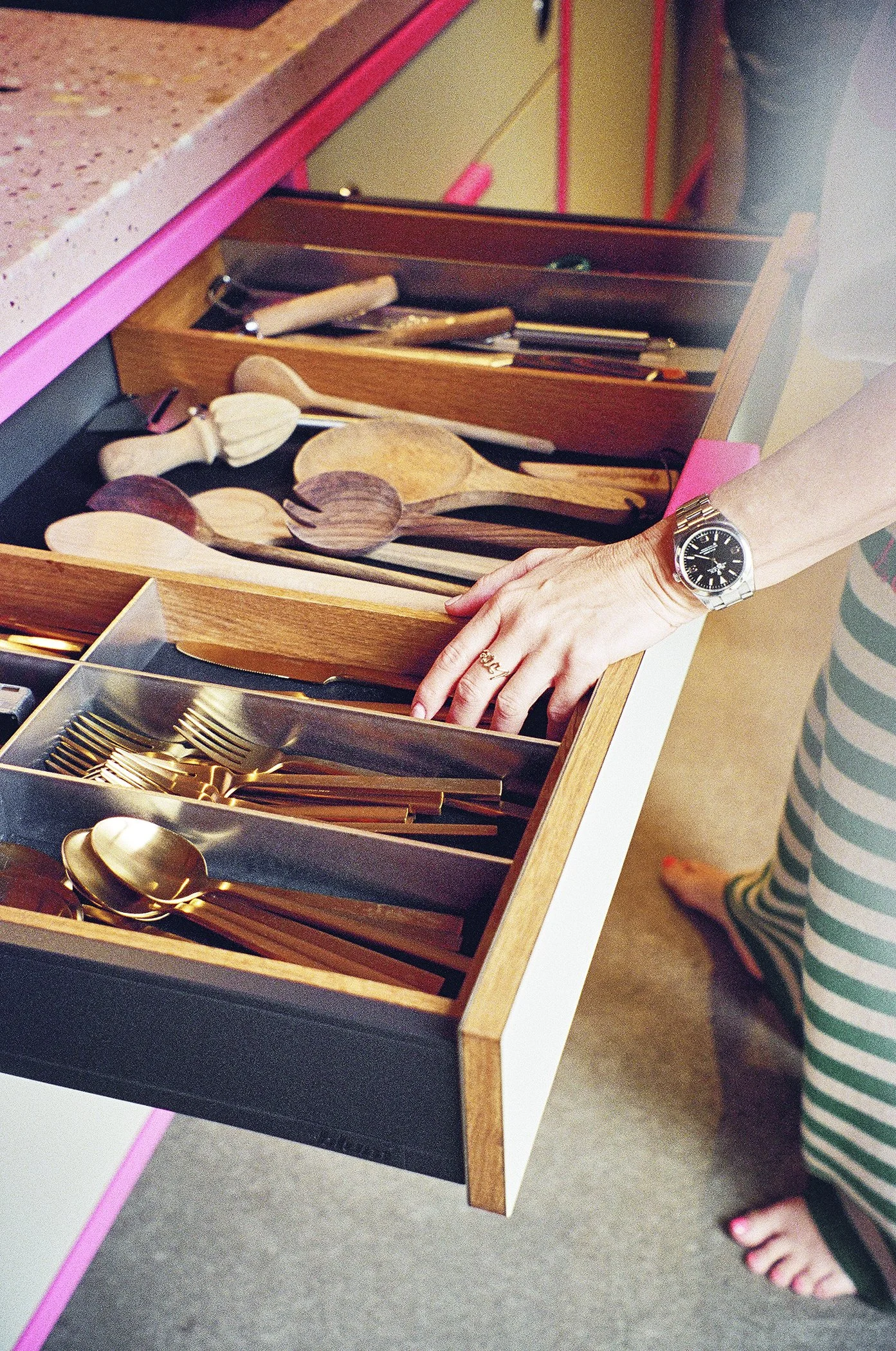 A person opening a drawer containing wooden spoons, gold utensils, and small kitchen tools, with some items organized in the sections.