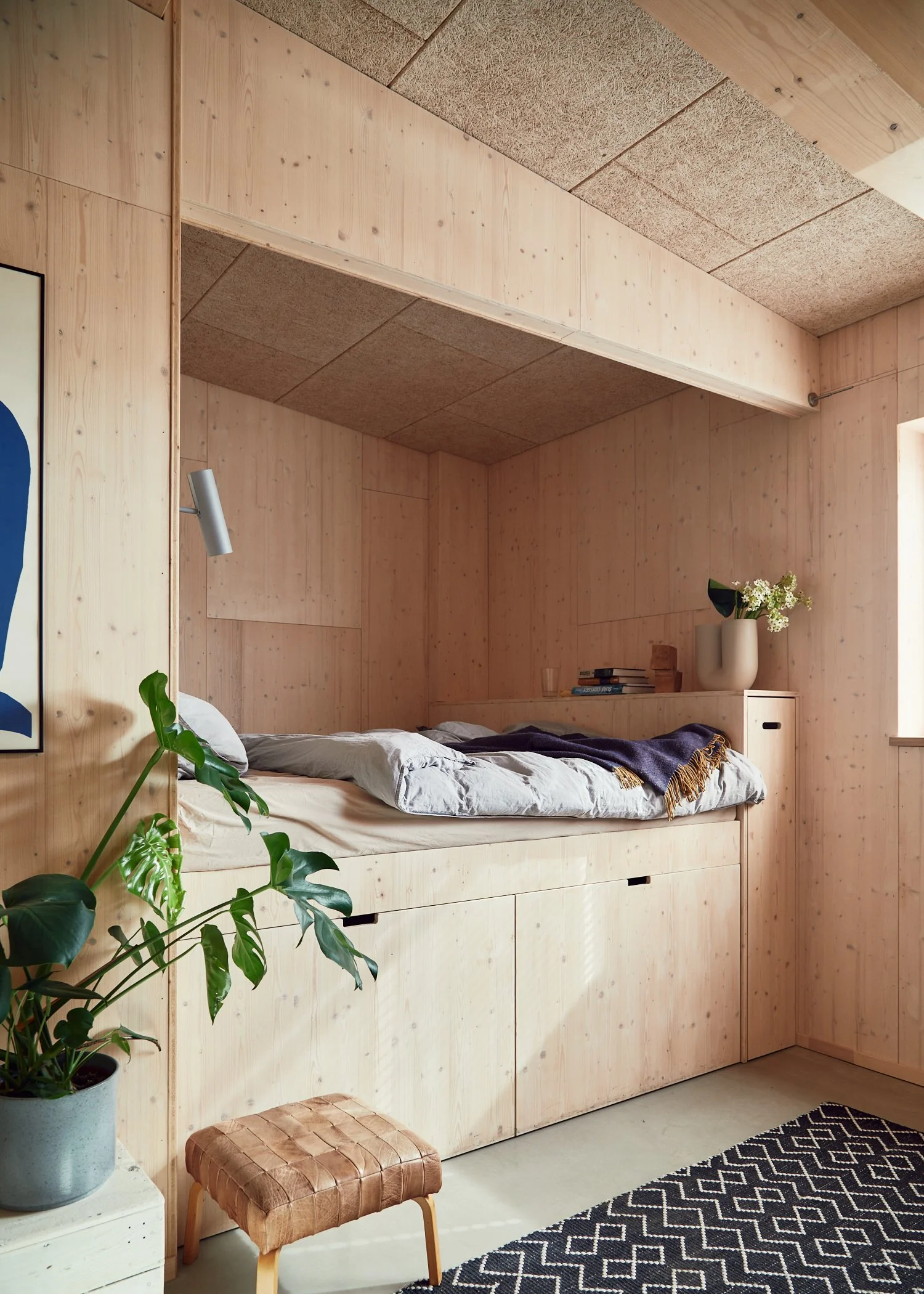 A bedroom with wooden paneling, a daybed with gray bedding, a large green plant in a pot, a small woven stool, and a black and white patterned rug.