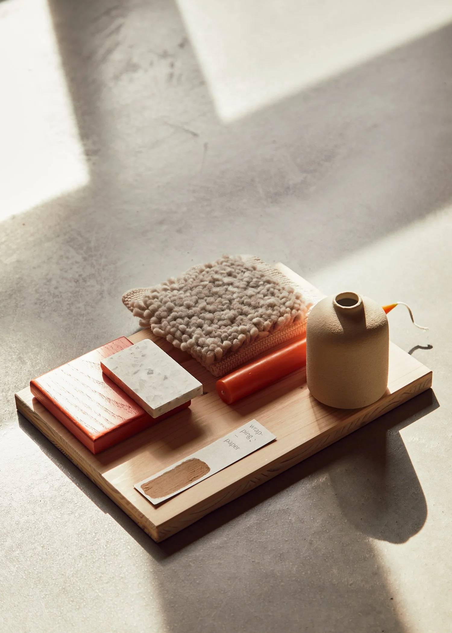 A wooden tray holding various textured and colored objects, including a small fluffy cloth, a rectangular marble piece, an orange slice, a beige vase, a small card with words, and a stick, with sunlight casting shadows on the surface.