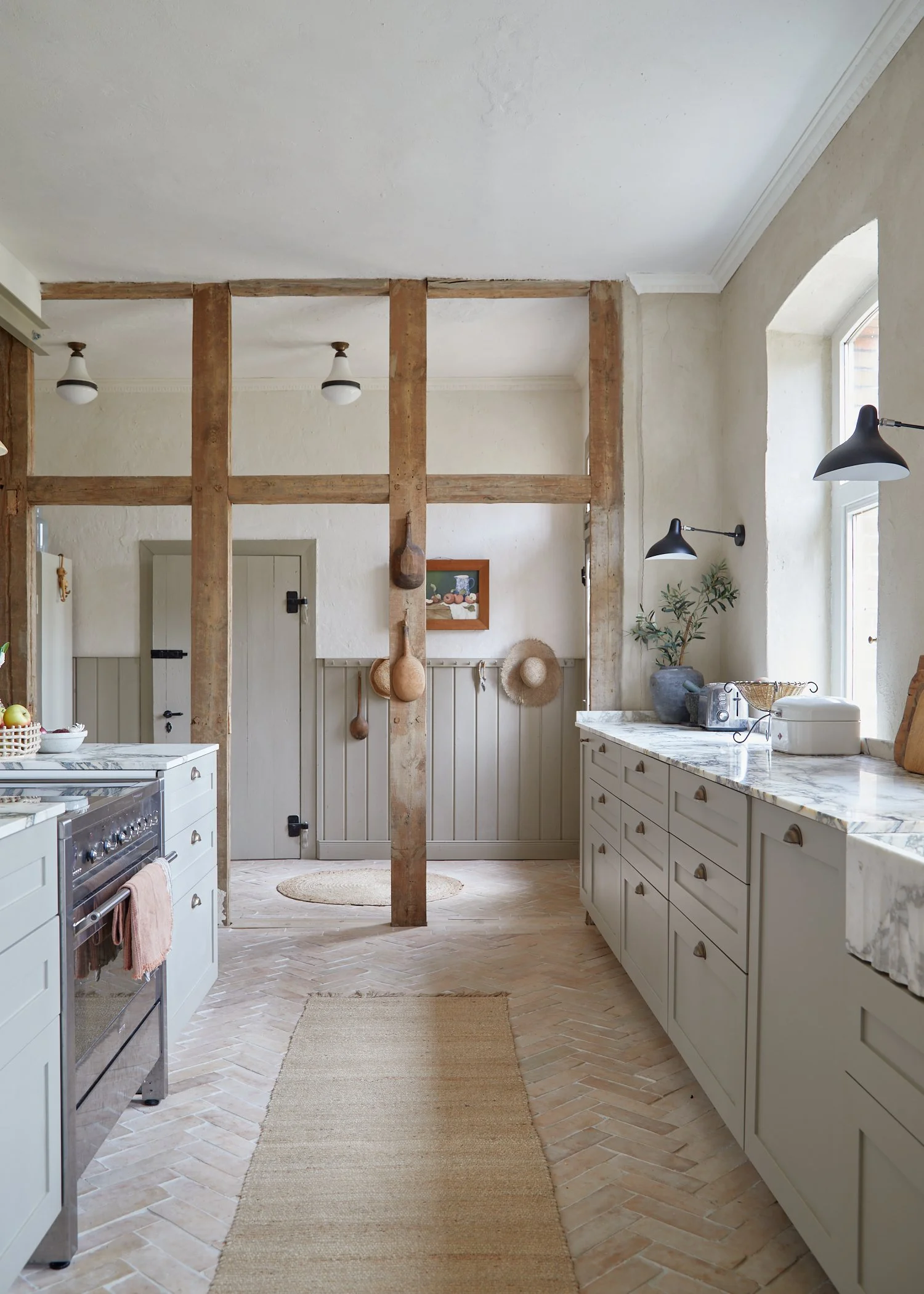 A rustic kitchen with white cabinets, marble countertops, and wooden beams, featuring hanging pots, hats, and decorative items, illuminated by natural light through the windows.