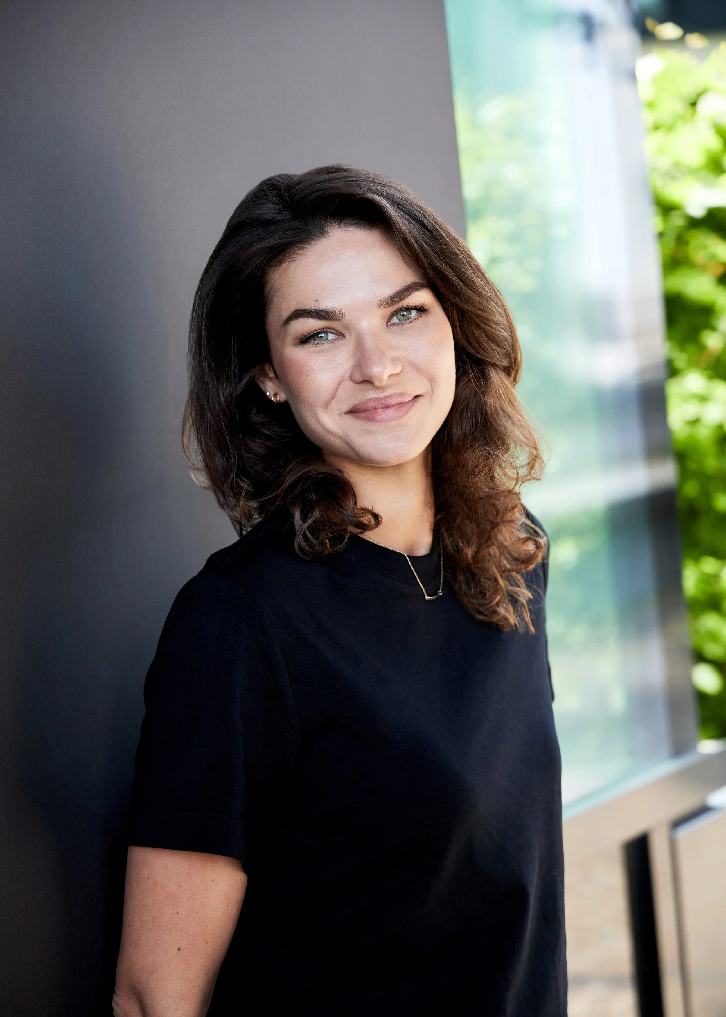 A woman with shoulder-length dark brown hair, light eyes, and fair skin is standing indoors against a dark wall and a large window with greenery outside. She is smiling and wearing a black T-shirt and a gold necklace.