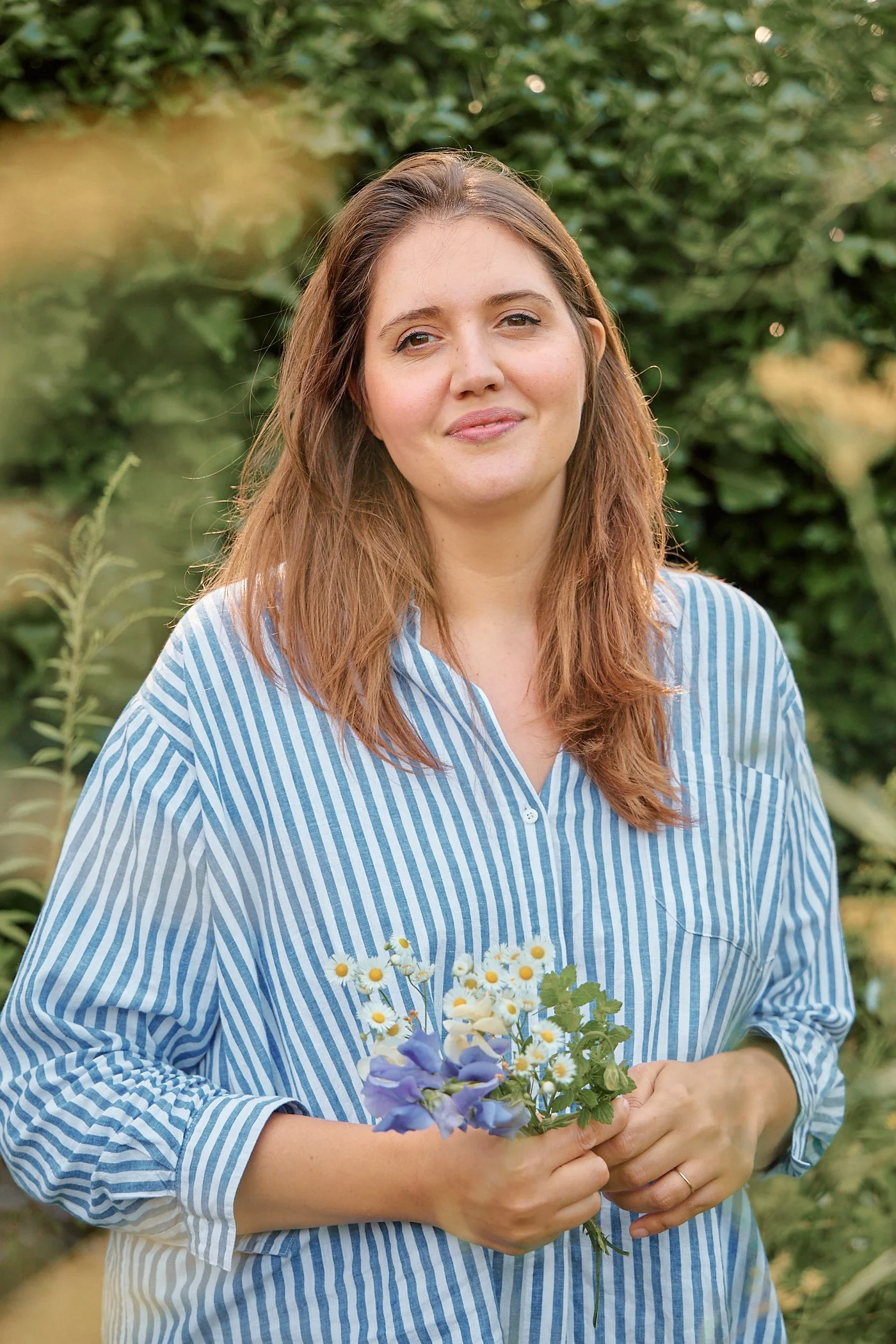 A woman with long, wavy, light brown hair holding a small bouquet of daisies and purple flowers outdoors with green foliage in the background.