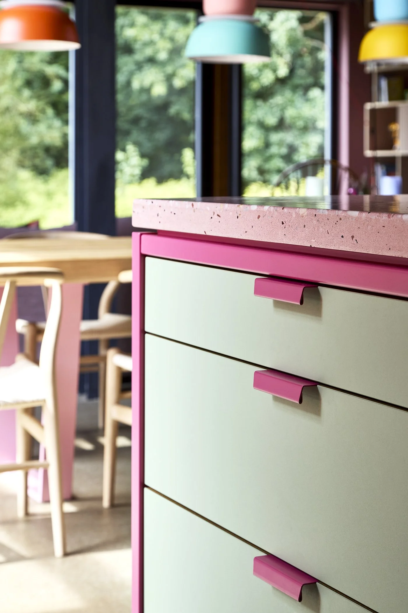 Close-up of a pastel green and pink dresser with pink handles in a bright, colorful room with wooden chairs and large windows showing greenery outside.