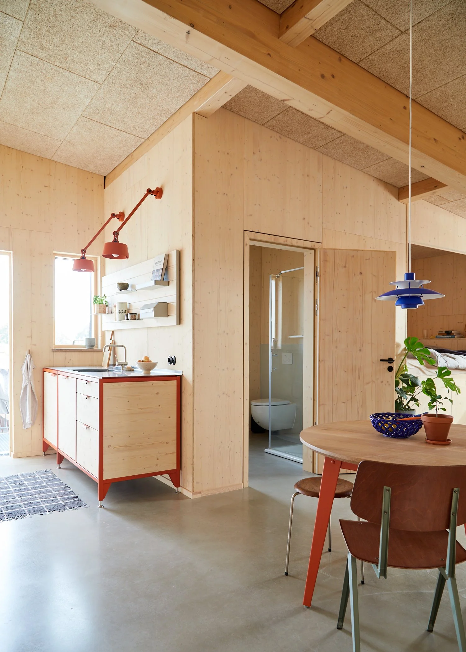 Interior of a modern minimalist wooden kitchen with a small dining table, a partial bathroom with a toilet and shower, and a decorative plant.