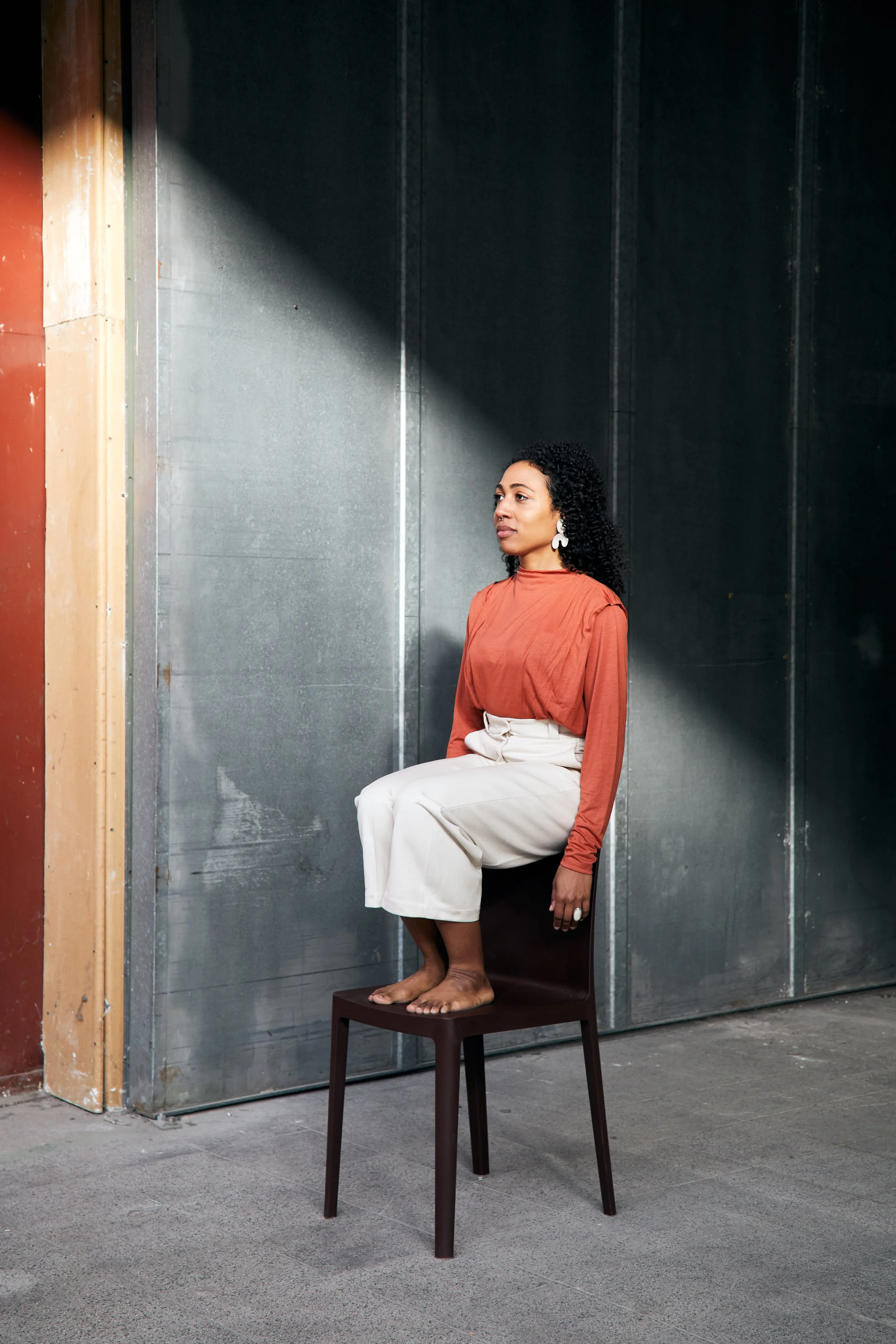 A woman sitting on a chair with her feet on another chair, against a metallic wall and industrial setting.