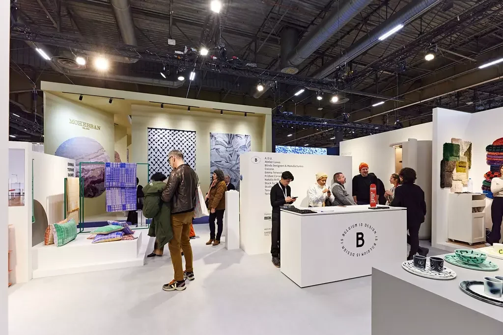 Exposition d'objets design et textiles dans un hall d'exposition, avec des visiteurs et un stand d'accueil.