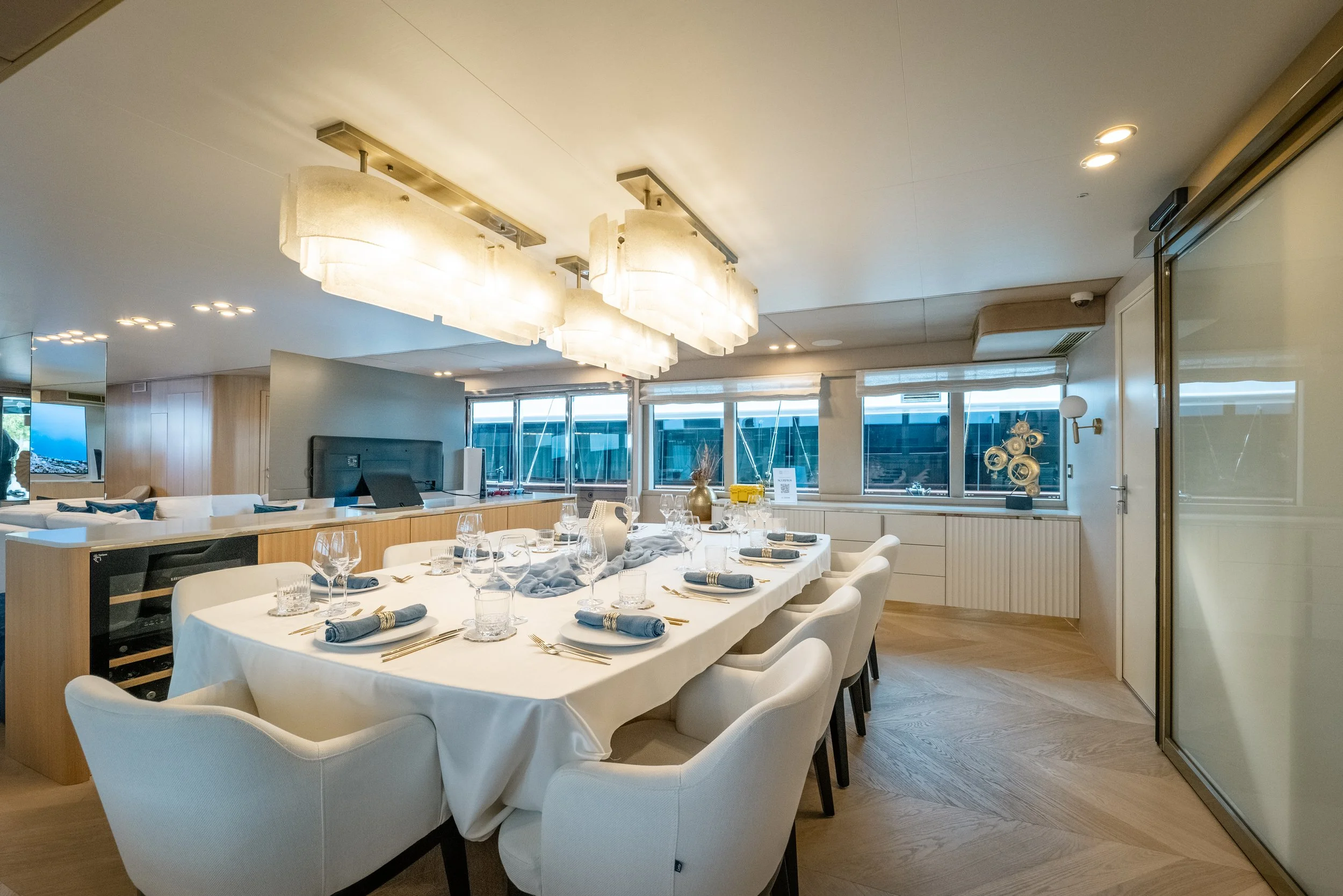 Elegant dining room with a long table set for a meal, featuring white tablecloths, blue napkins, glassware, and gold utensils, with modern lighting fixtures and large windows.