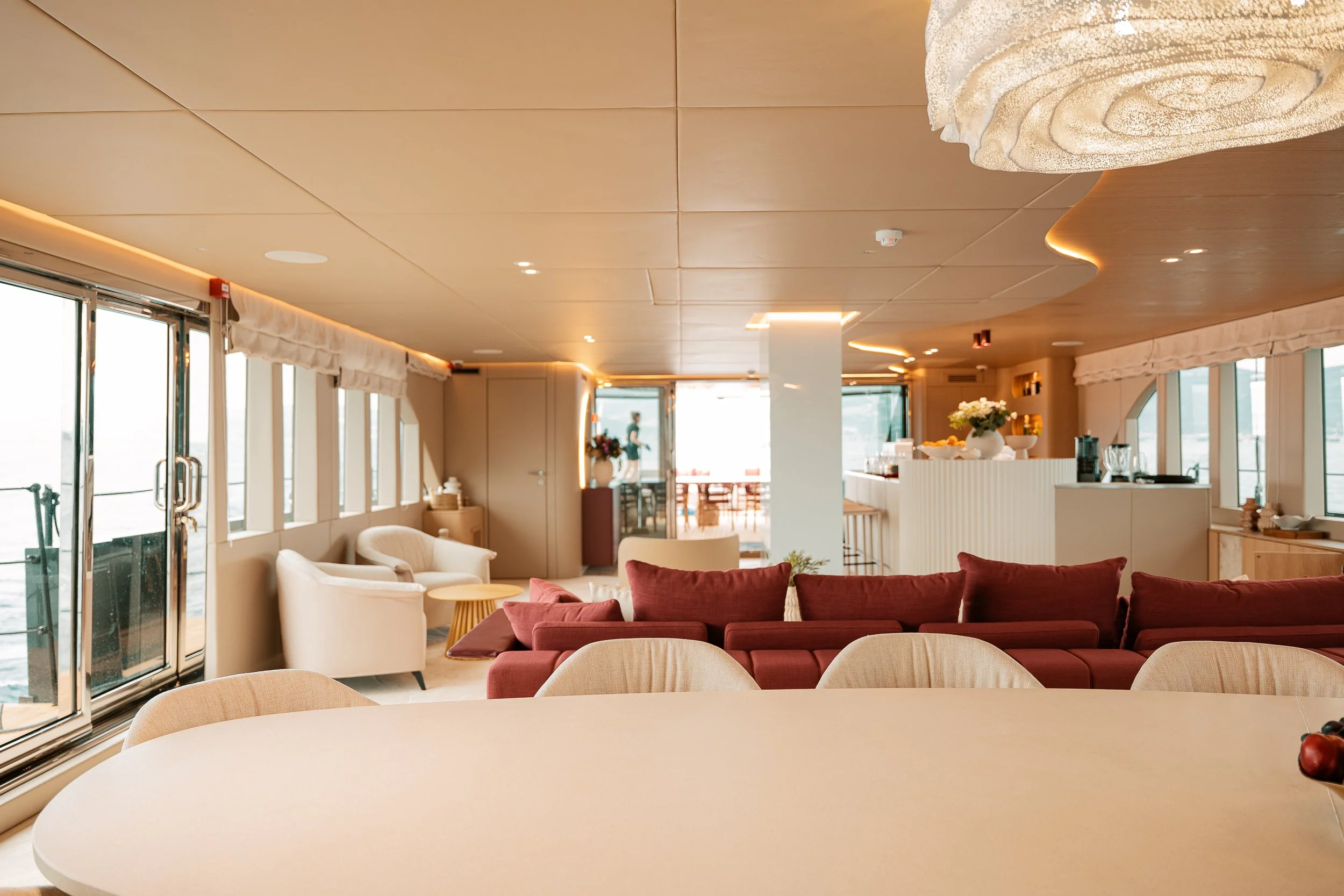 Interior of a spacious, well-lit lounge with large windows, red and beige sofas, white armchairs, a bar area with flowers, and a dining area in the background.