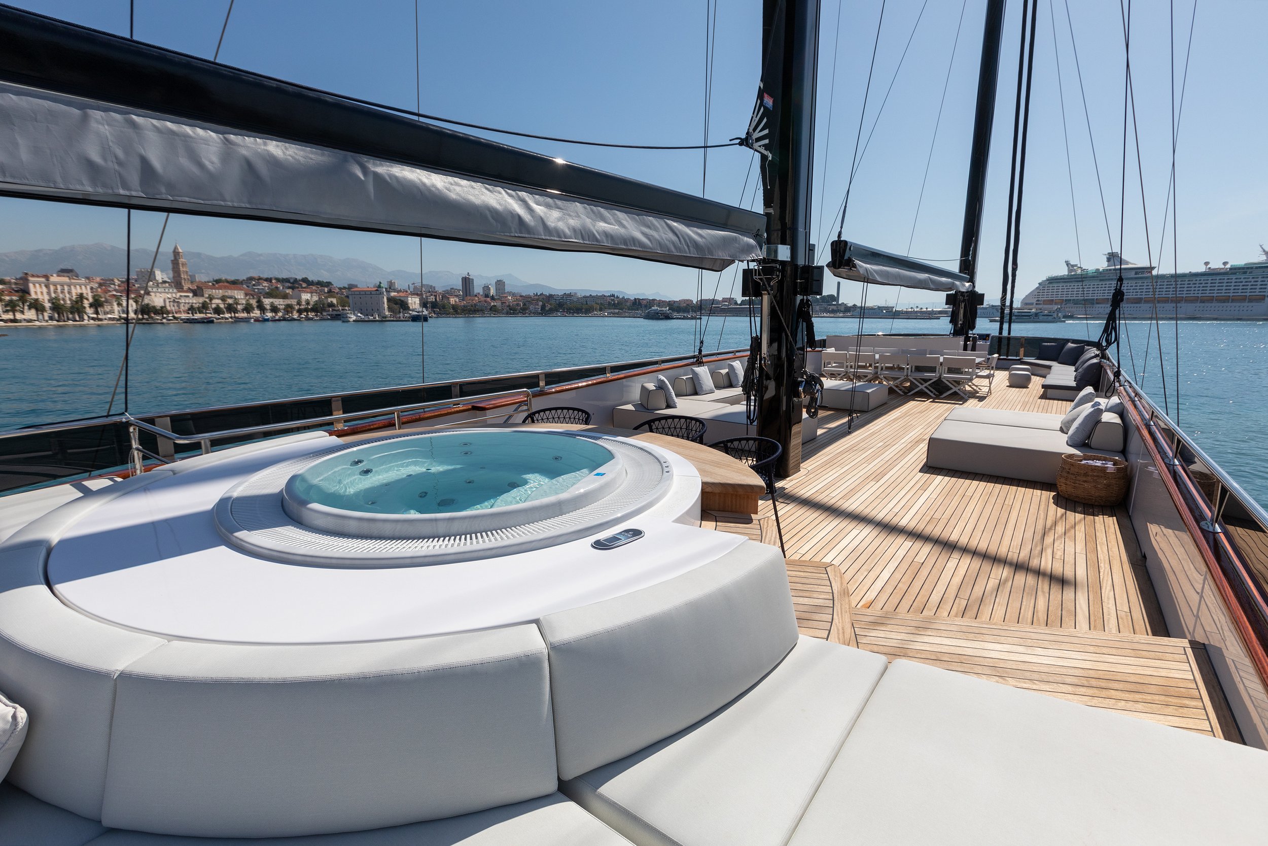 Luxury yacht deck with a hot tub, cushioned seating, and a scenic city waterfront view in the background.