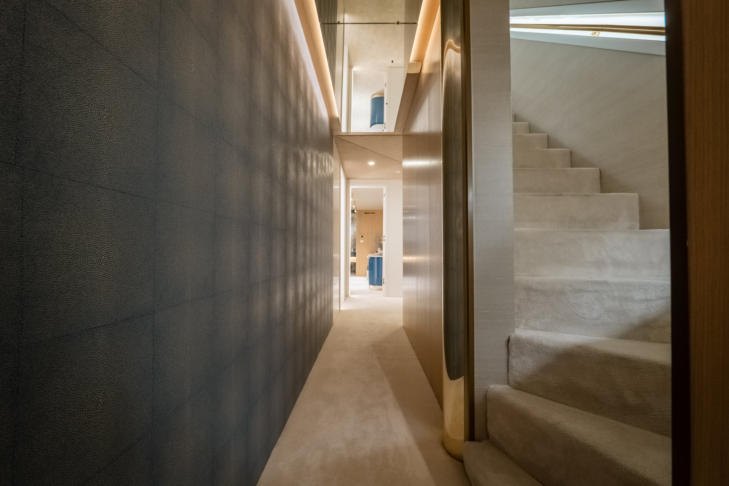 View of a modern, well-lit hallway with stairs on the right side, and a door at the end leading to a bright room, featuring neutral tones and minimalist design.
