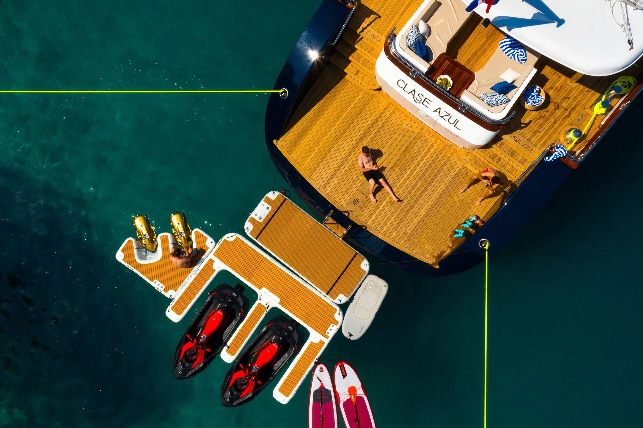 Overhead view of a yacht with a wooden deck labeled 'Clase Azul,' two jet skis, a paddleboard, and three people relaxing or preparing water activities on a clear blue body of water.