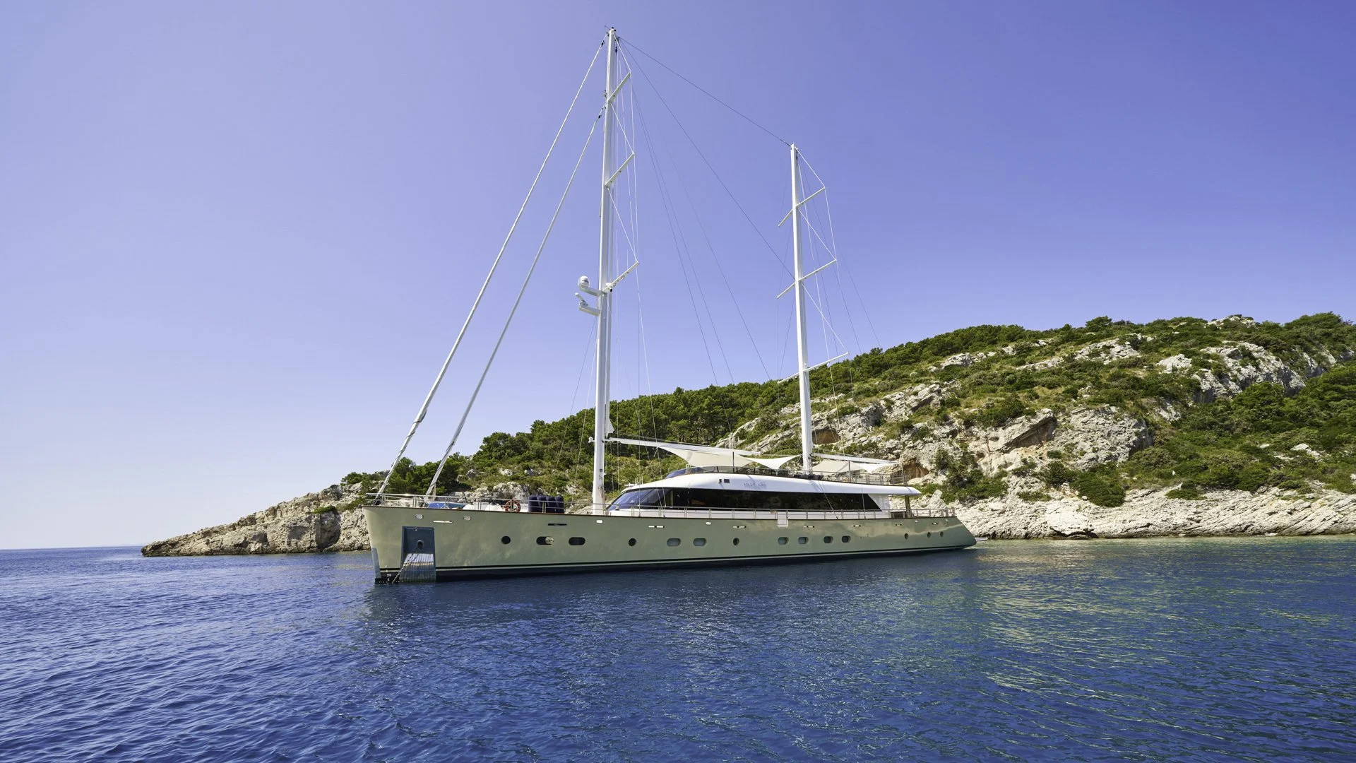 A large white sailing yacht with two masts anchored near a rocky, green hillside on a clear day, with calm blue water surrounding it.