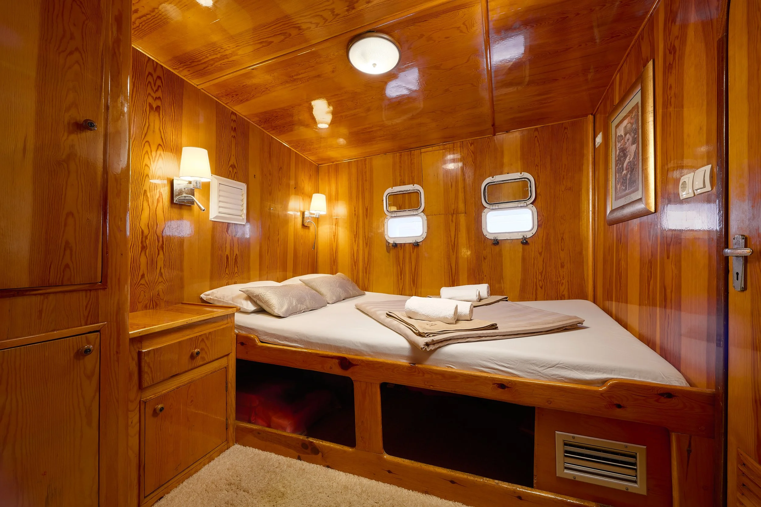 Wood-paneled cabin bedroom with a large bed, two small windows, wall-mounted lamps, framed artwork, and a vent near the floor.