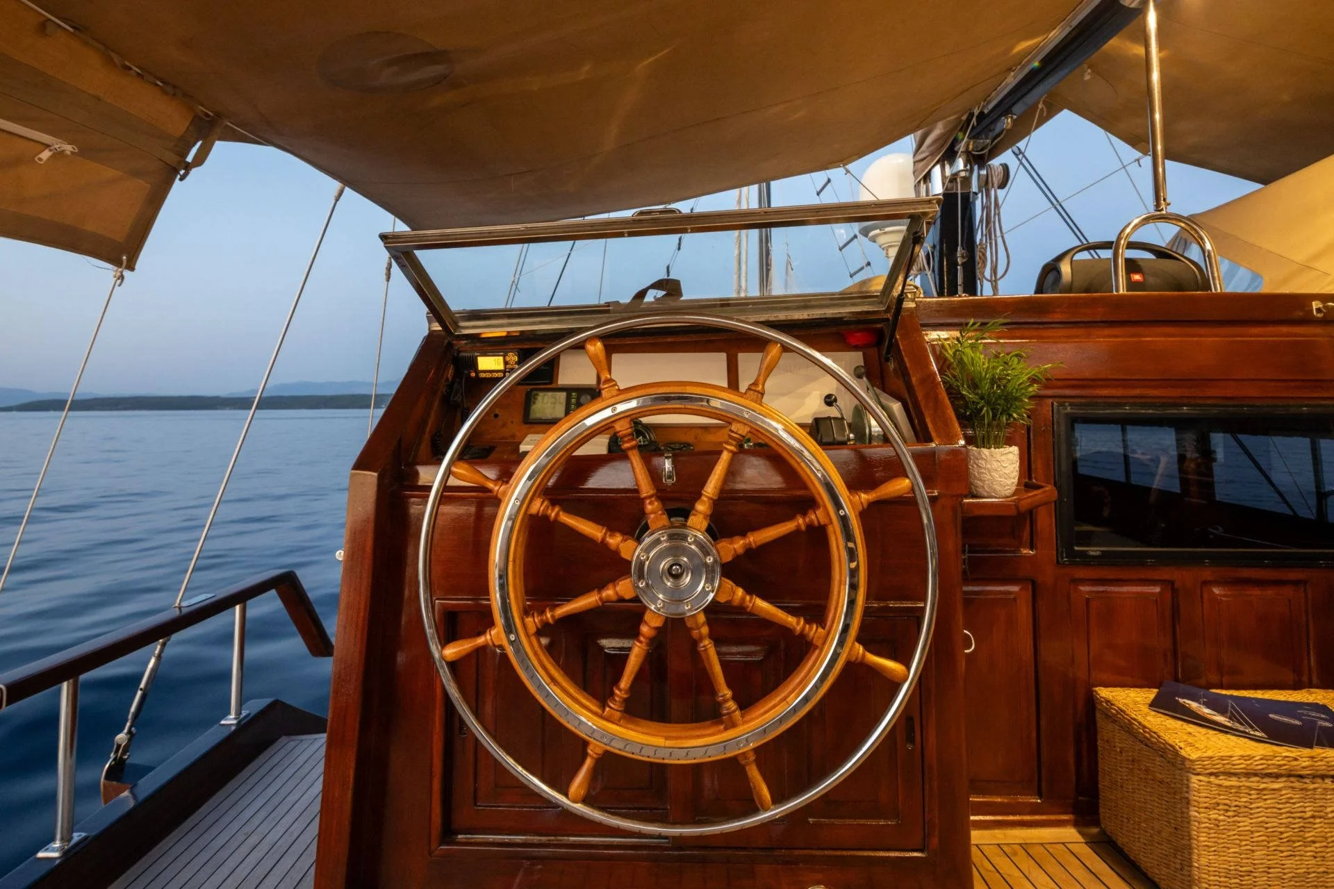 Wooden sailboat steering wheel with nautical instruments and a potted plant, set against a calm blue sea and wooden deck.
