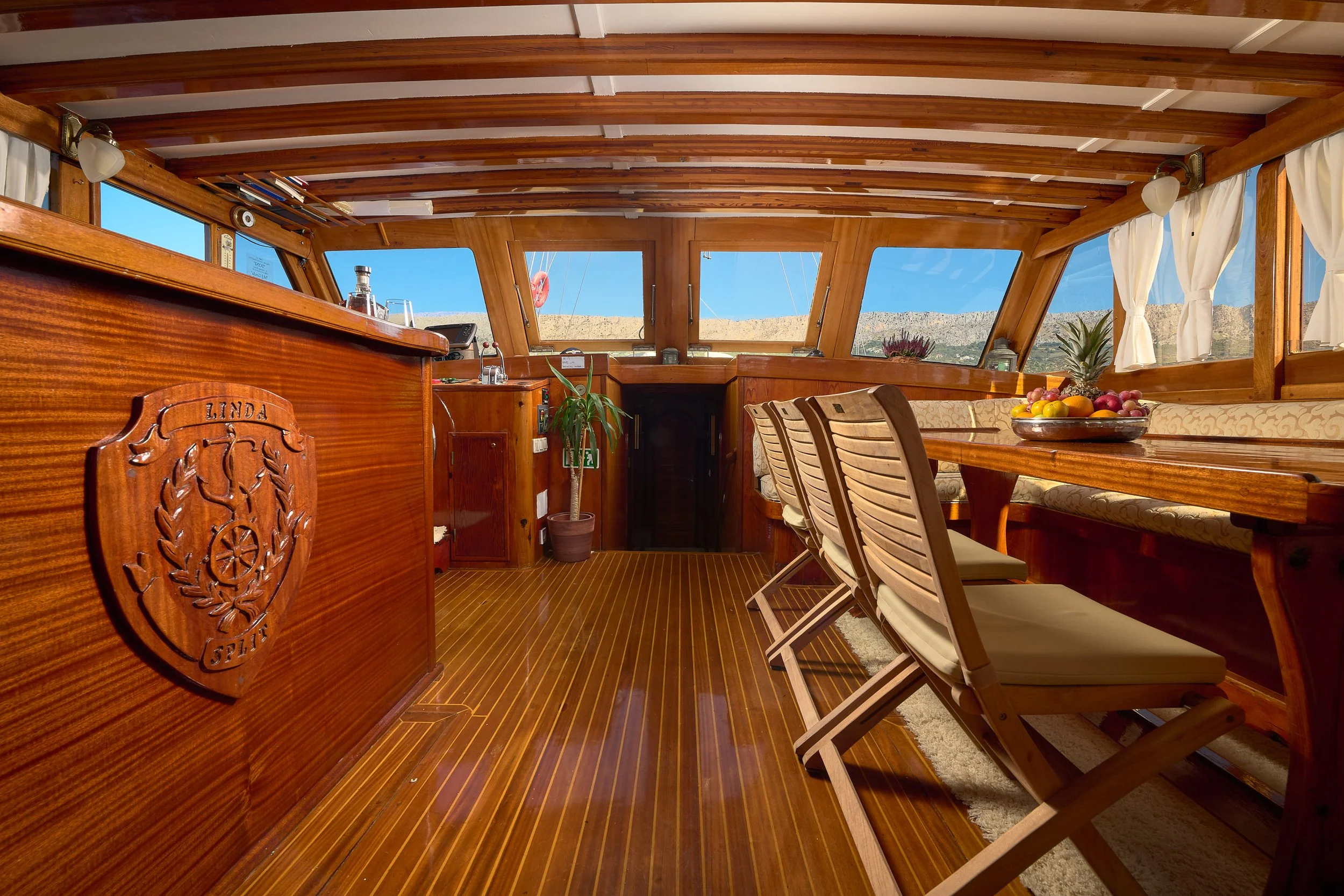 The interior of a wooden boat's cabin featuring a polished wooden bar with a crest that reads "Linda 717," a table with four chairs, windows with white curtains, and a view of blue sky and distant mountains.