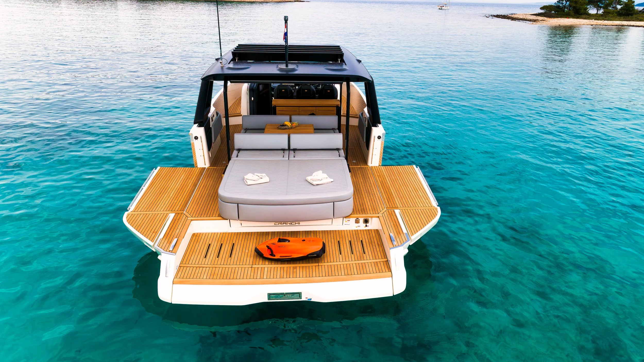 A luxury yacht with a spacious wooden deck floats on clear blue water. The deck features a cushioned sunbathing area, a table, and some towels. There is an orange remote control or device on the deck. The yacht's interior and a distant shoreline are 
