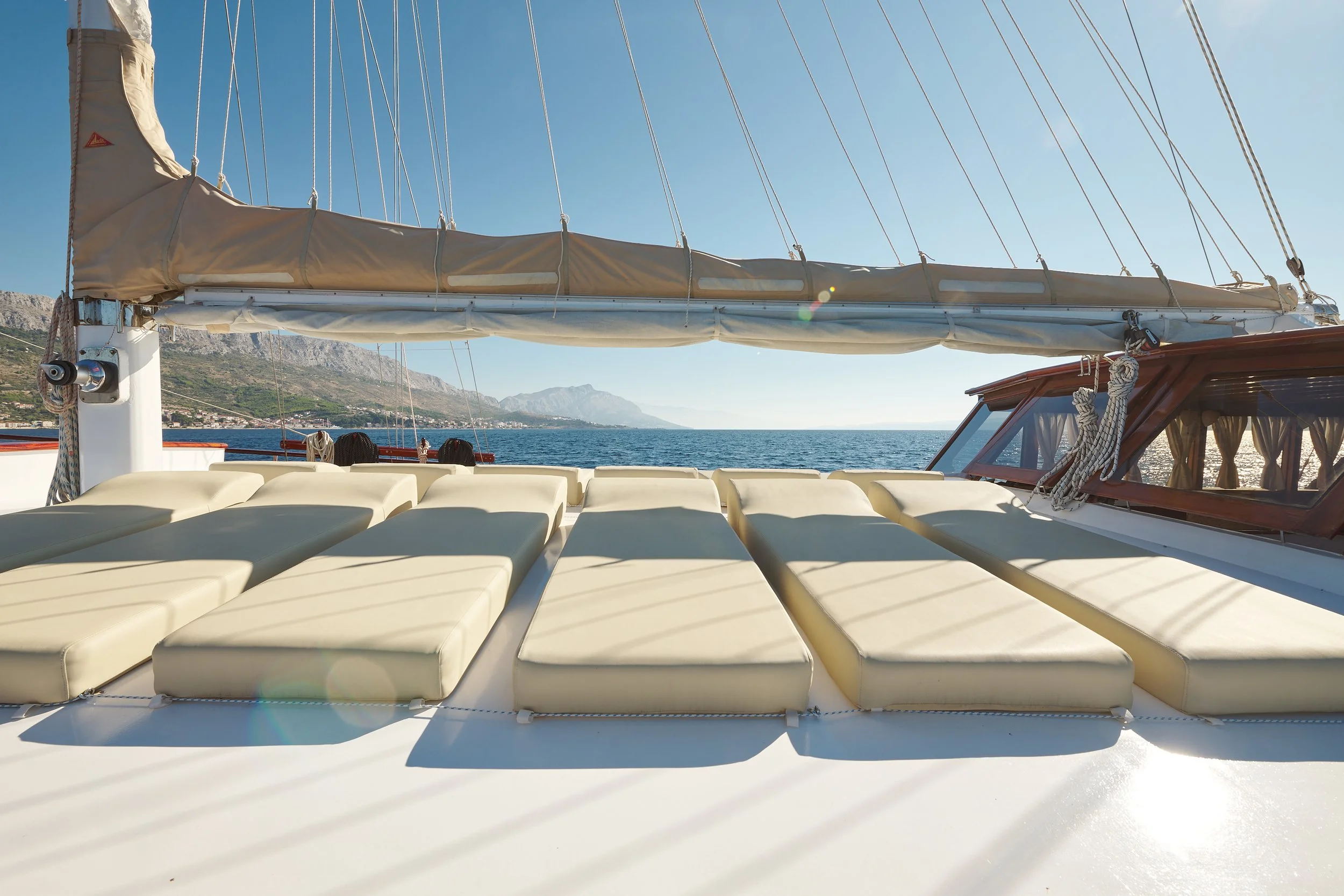 On a boat, beige cushions laid out on the deck, with a view of the blue sea, shoreline, and mountains in the distance under a clear sky.