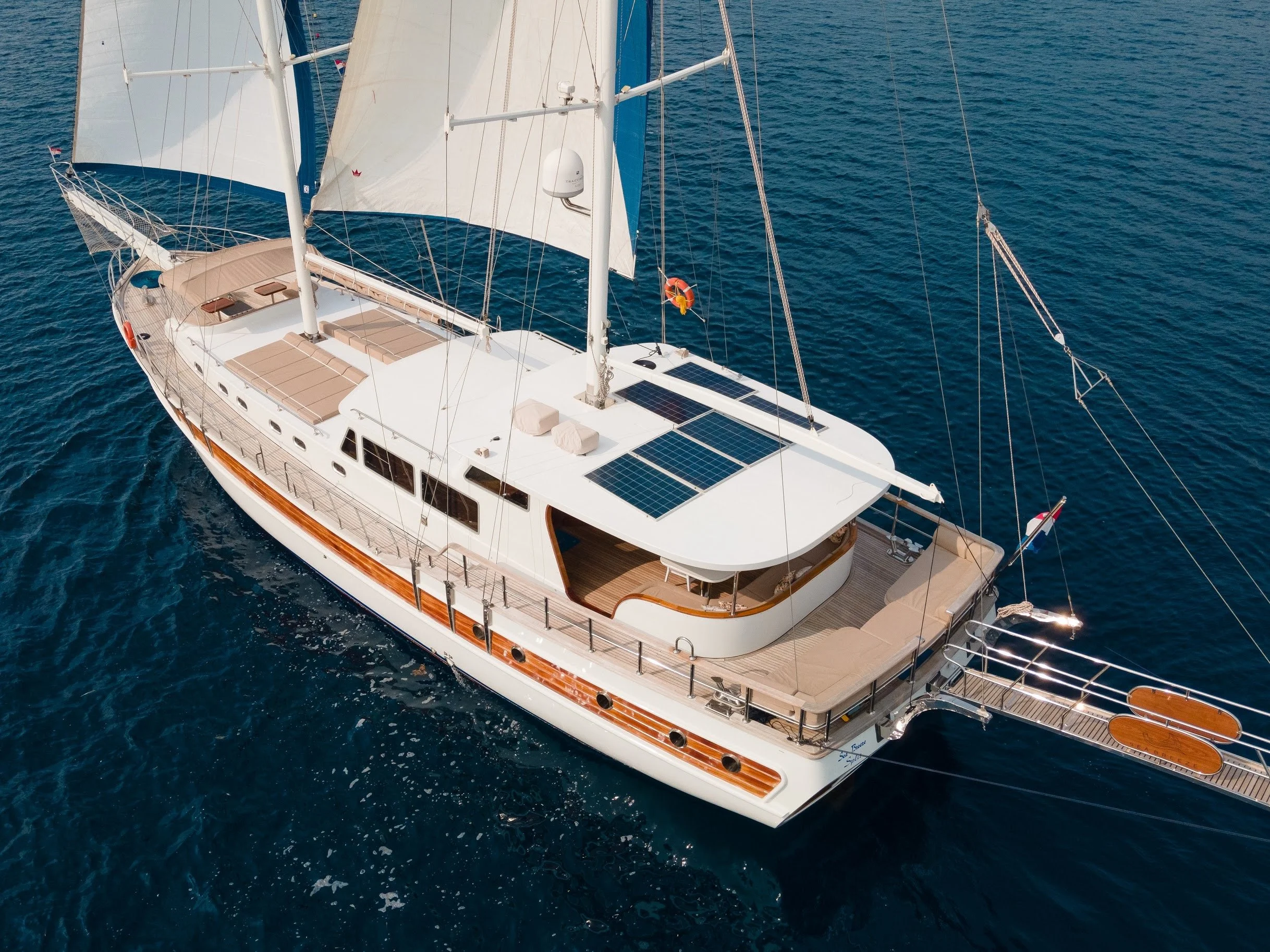 An aerial view of a white sailing yacht with brown accents anchored on deep blue water. The yacht has multiple sails, solar panels on the roof, and an open deck with lounge seating.