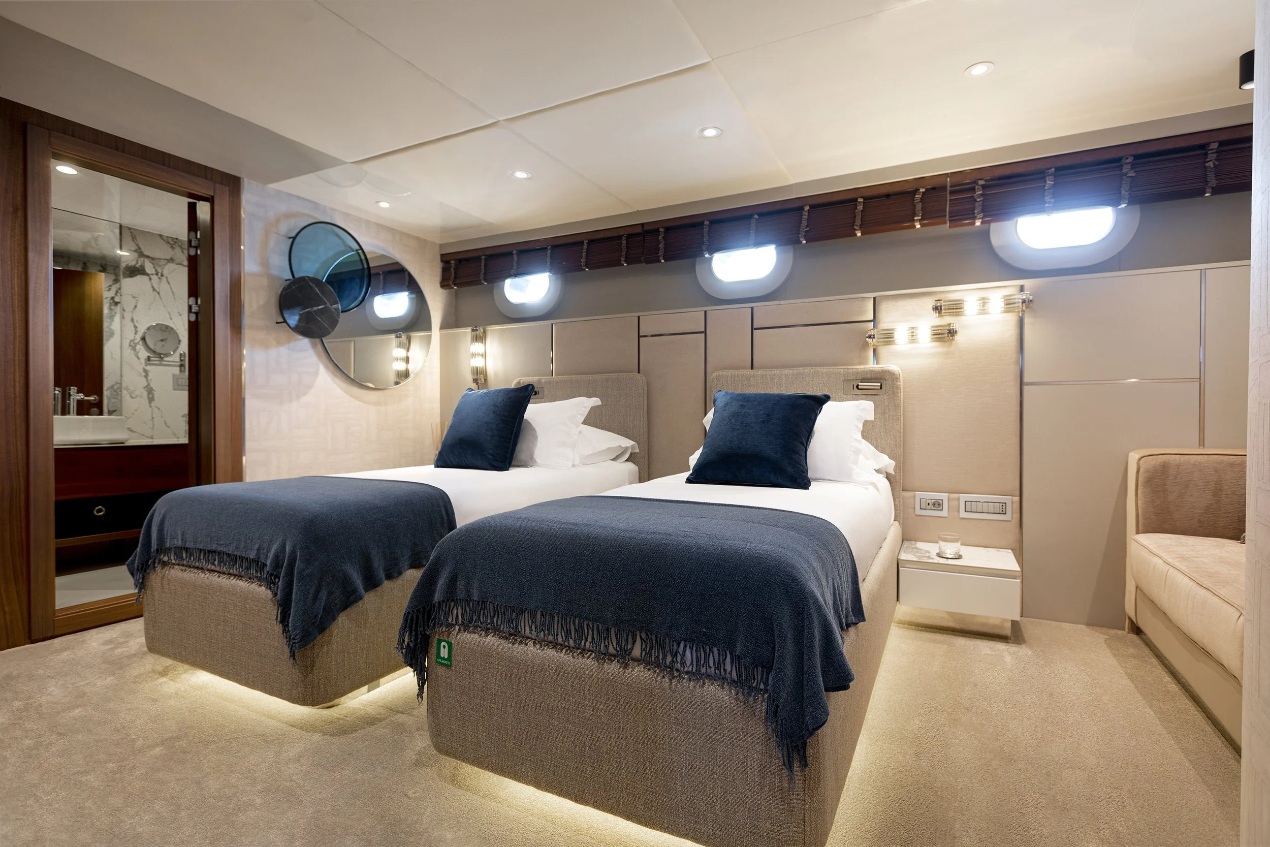 Hotel room with two beds, navy blue throws and pillows, beige headboards, wall-mounted lights, round mirror, sitting area, and bathroom visible through door.