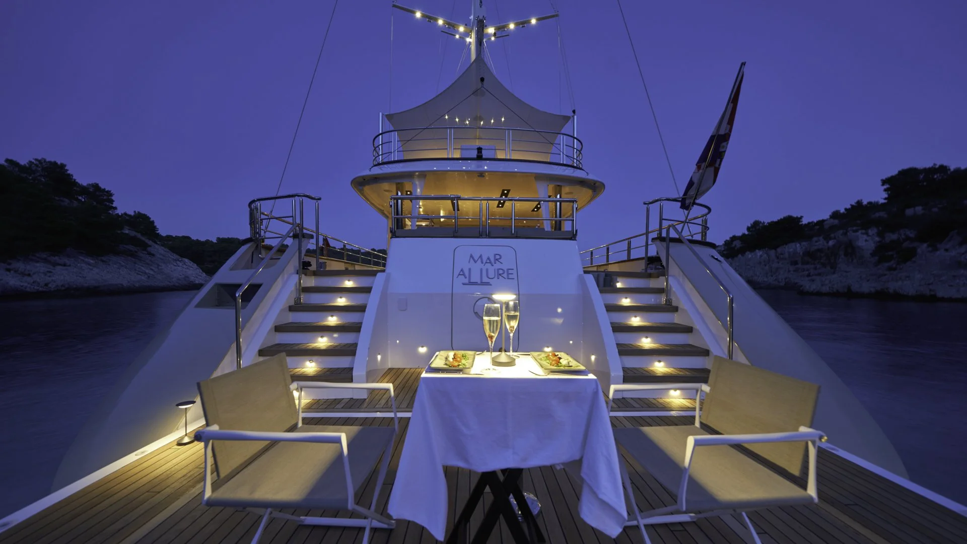 A yacht named 'Mar Allure' docked at dusk, with a table set for two on the deck, including two glasses of champagne, plates, and utensils, illuminated by small lights along the stairs and deck.