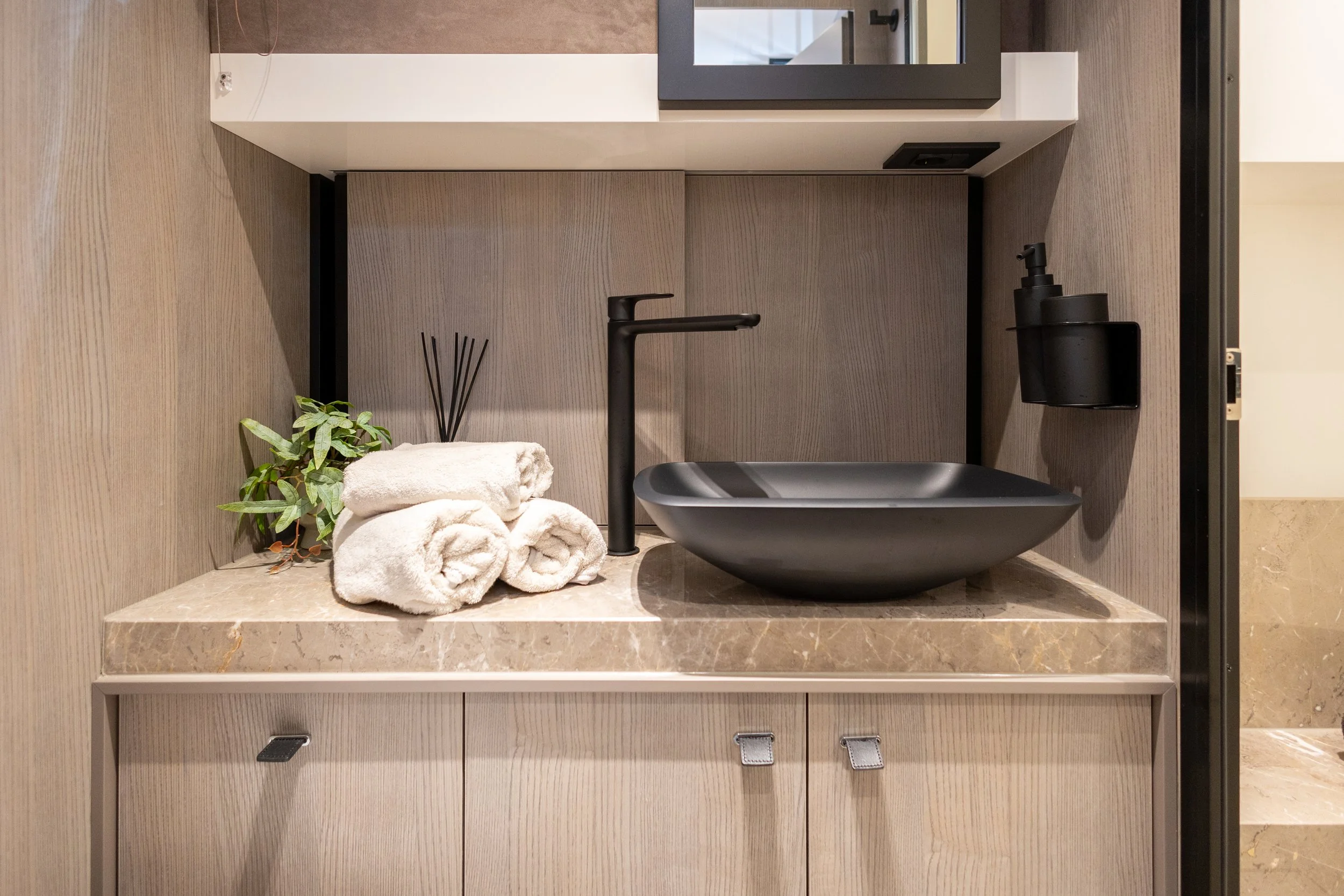 Bathroom sink with black basin, black faucet, rolled white towels, green plant, black soap dispenser, and reed diffuser on beige marble countertop with light wood cabinet below.