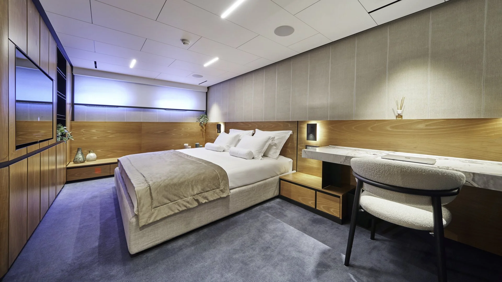 Modern hotel room with a bed, wooden paneling, a desk with a chair, and a TV on the left wall.