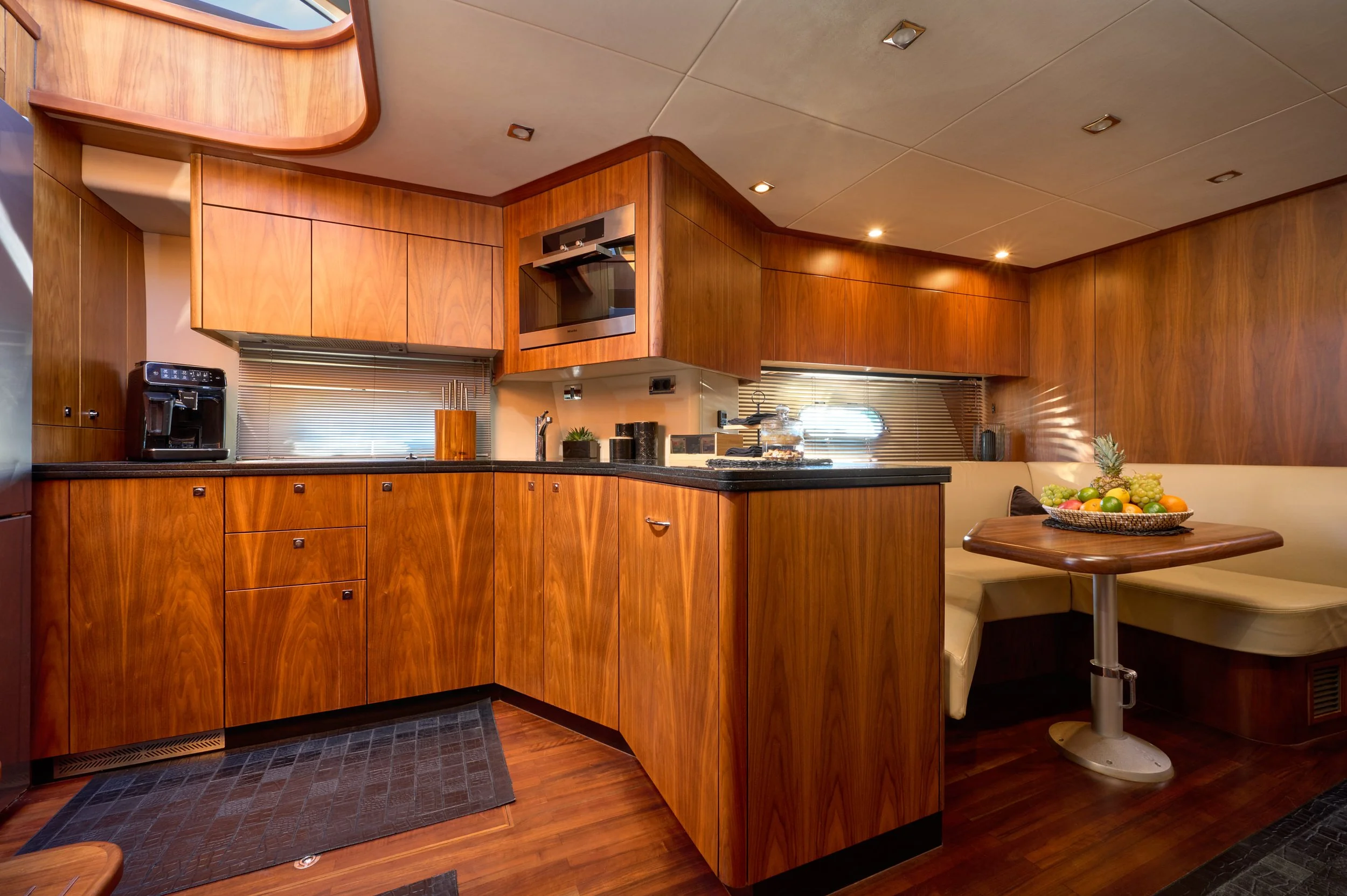 A kitchen with wood paneling, a black countertop, a coffee machine, a microwave oven, a small table with a fruit basket, and built-in cabinets and seating area.
