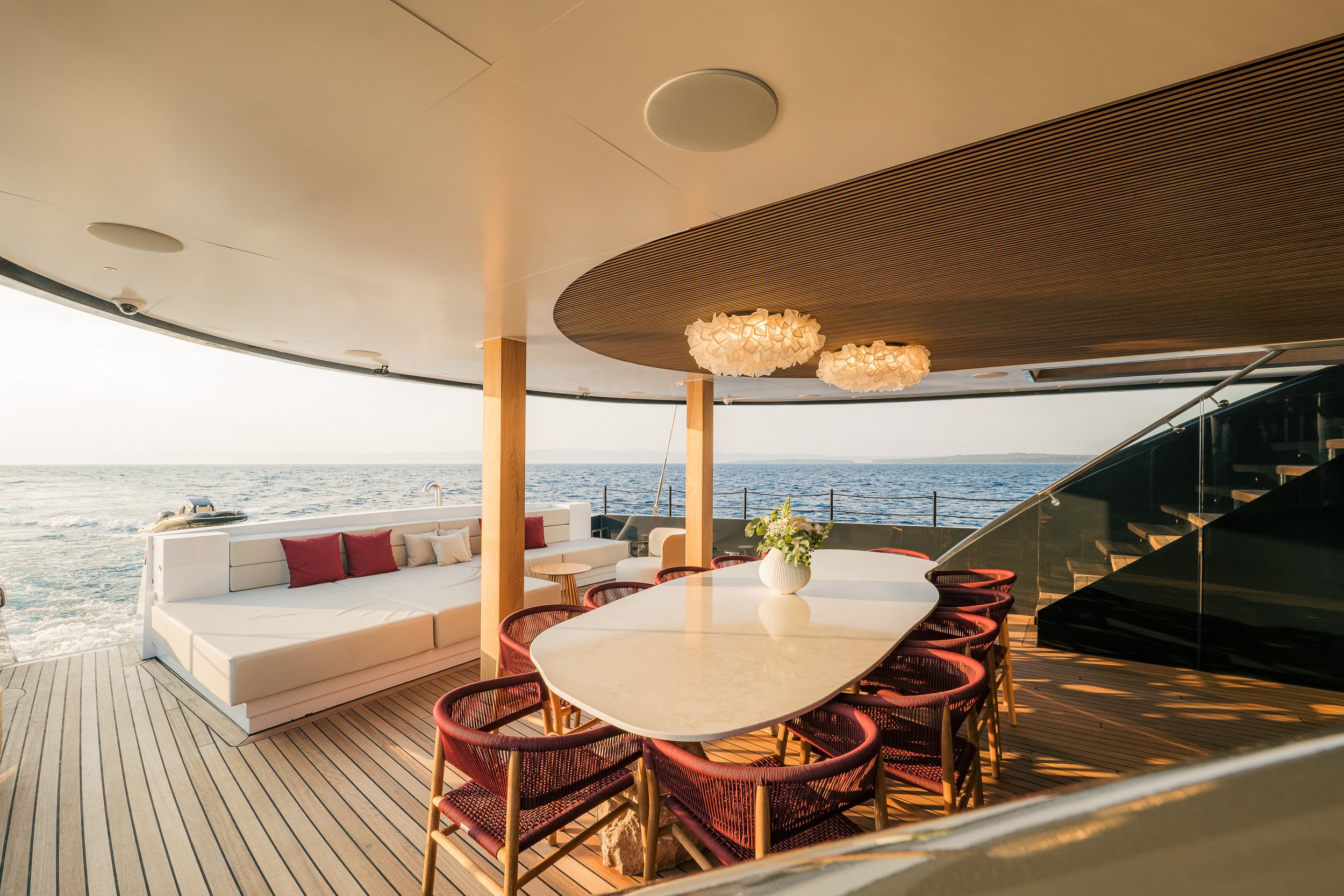 An outdoor dining and lounging area on a yacht with a view of the ocean, featuring a white sofa with red and beige pillows, a white oval table with a flower arrangement, red chairs, and wooden flooring.