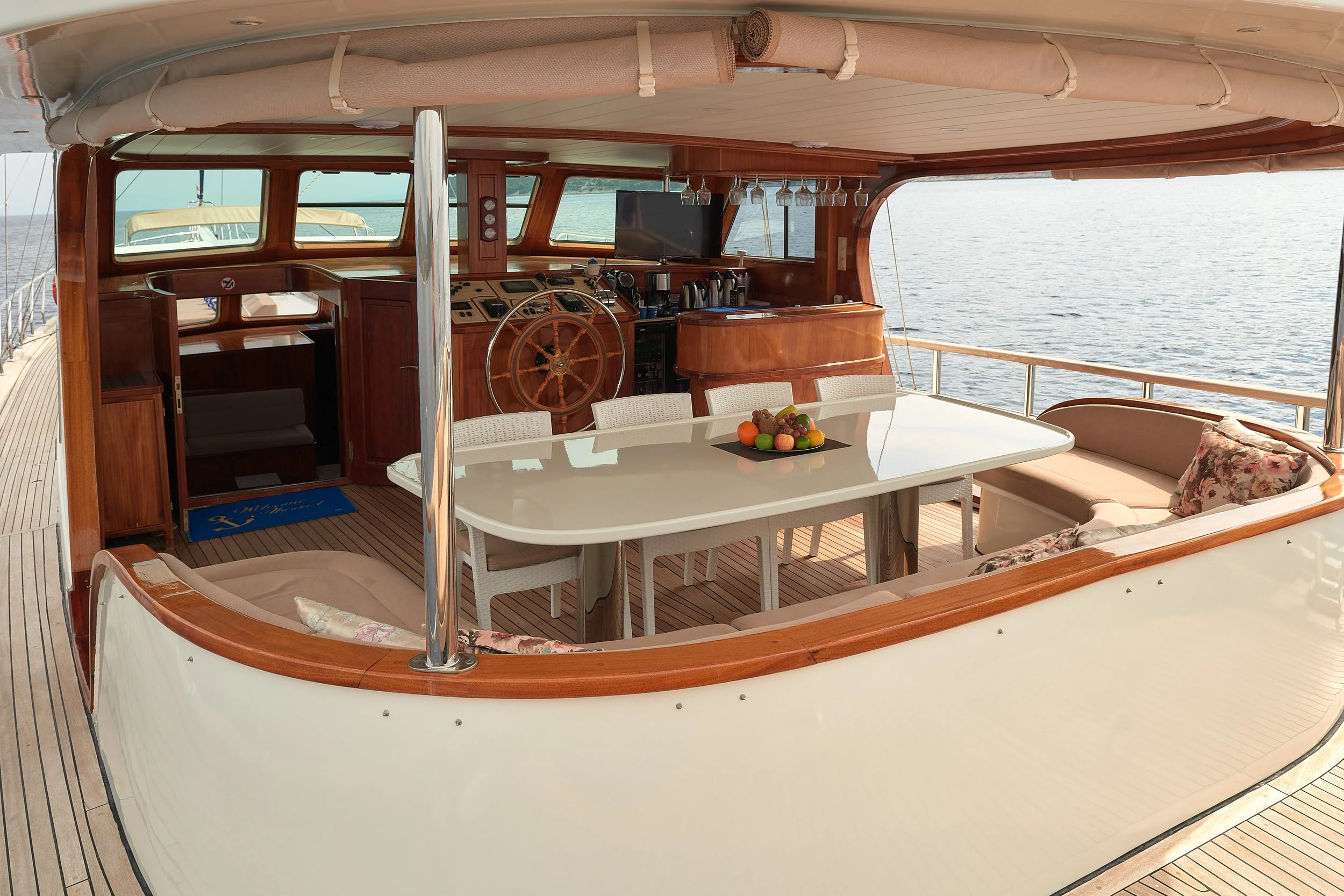 Outdoor deck seating area on a yacht with a large white table, cushioned seating, a fruit bowl, and a wooden helm station in the background with navigation equipment and a TV, overlooking water.