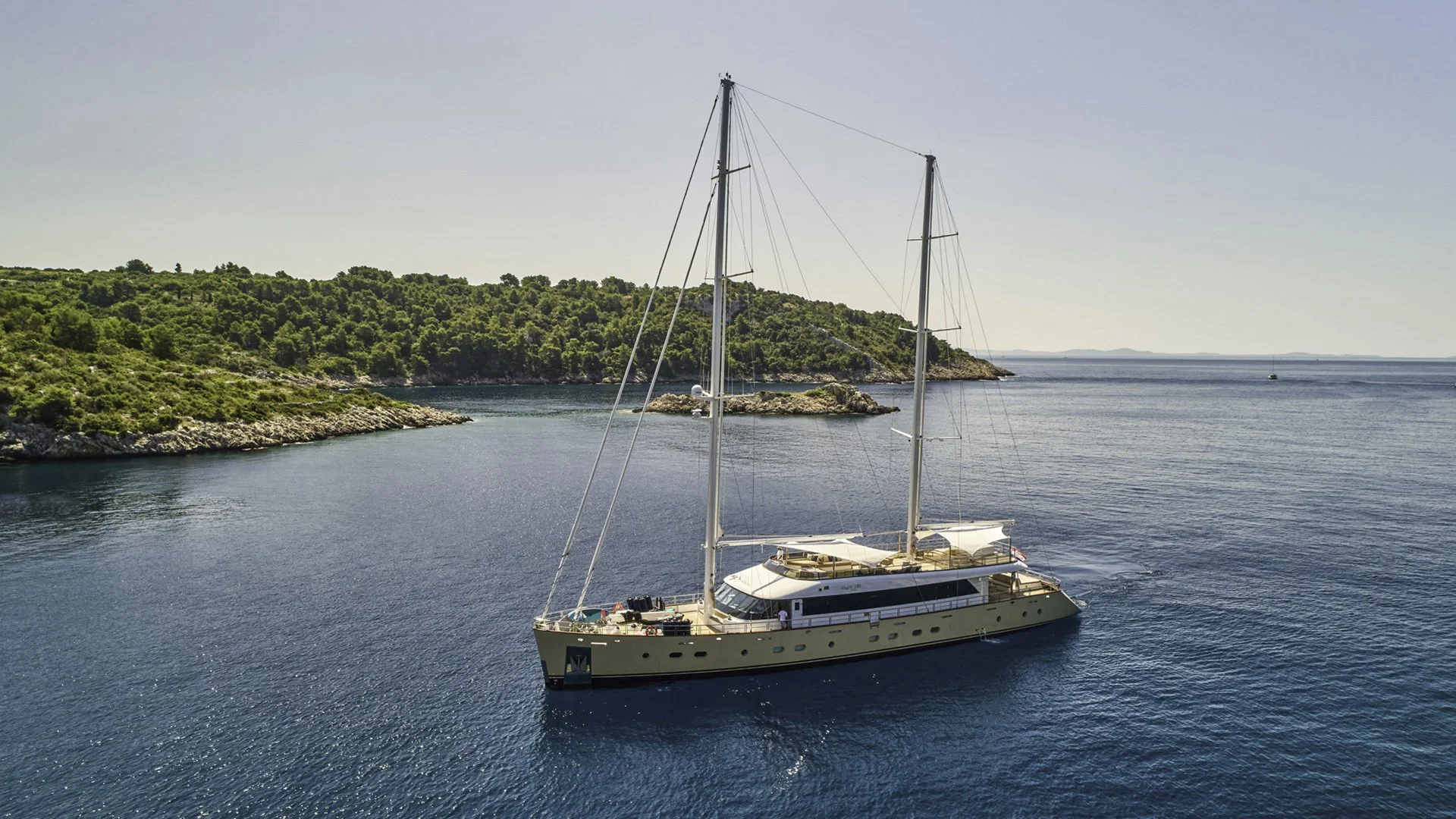 A large white sailing yacht with two masts floating on calm blue water near a green, rocky coastline with trees and bushes.