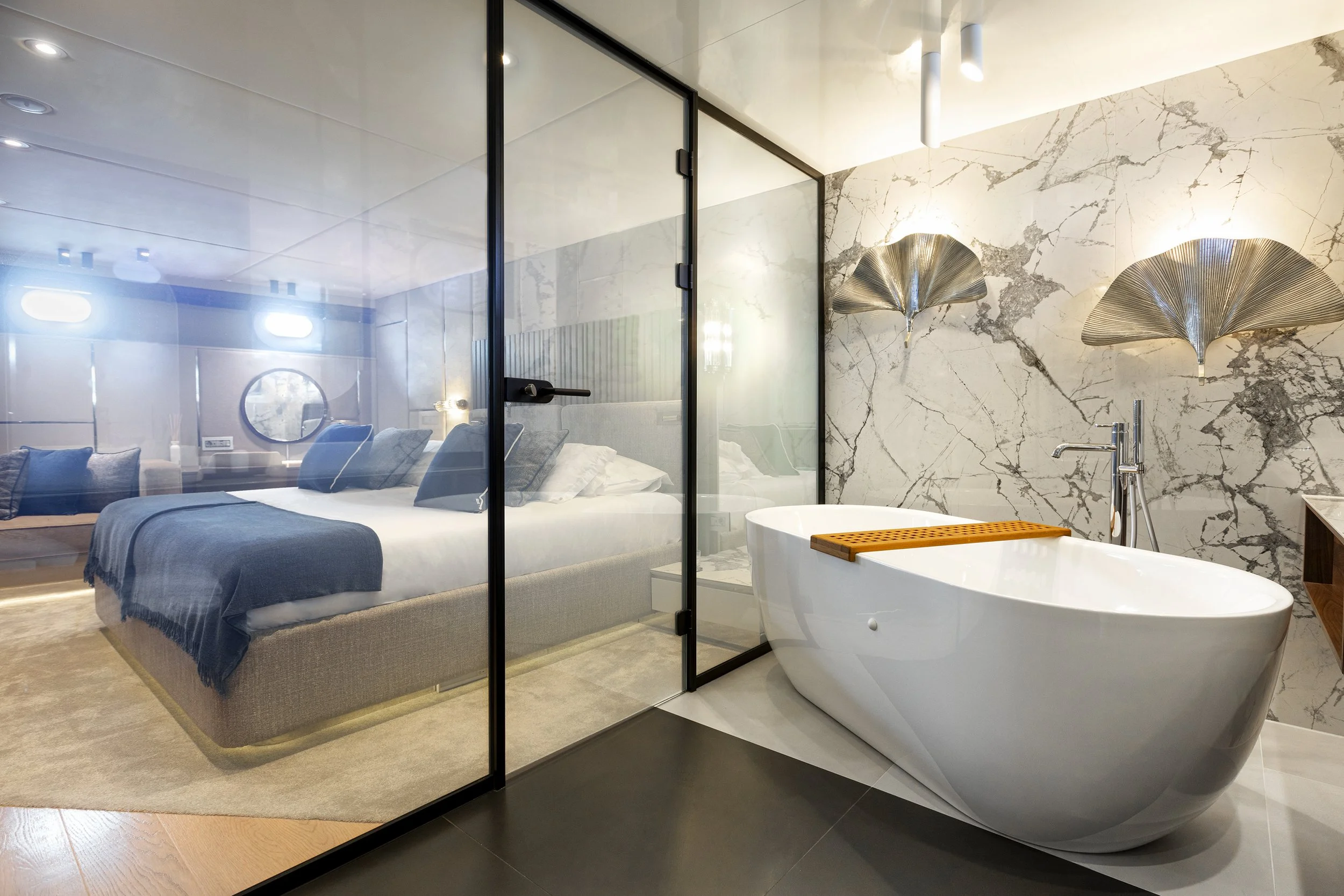 Modern hotel room with a glass wall separating a bedroom with a bed and a sitting area, and a bathroom with a freestanding bathtub against a marble wall decorated with metallic fan-shaped wall art.